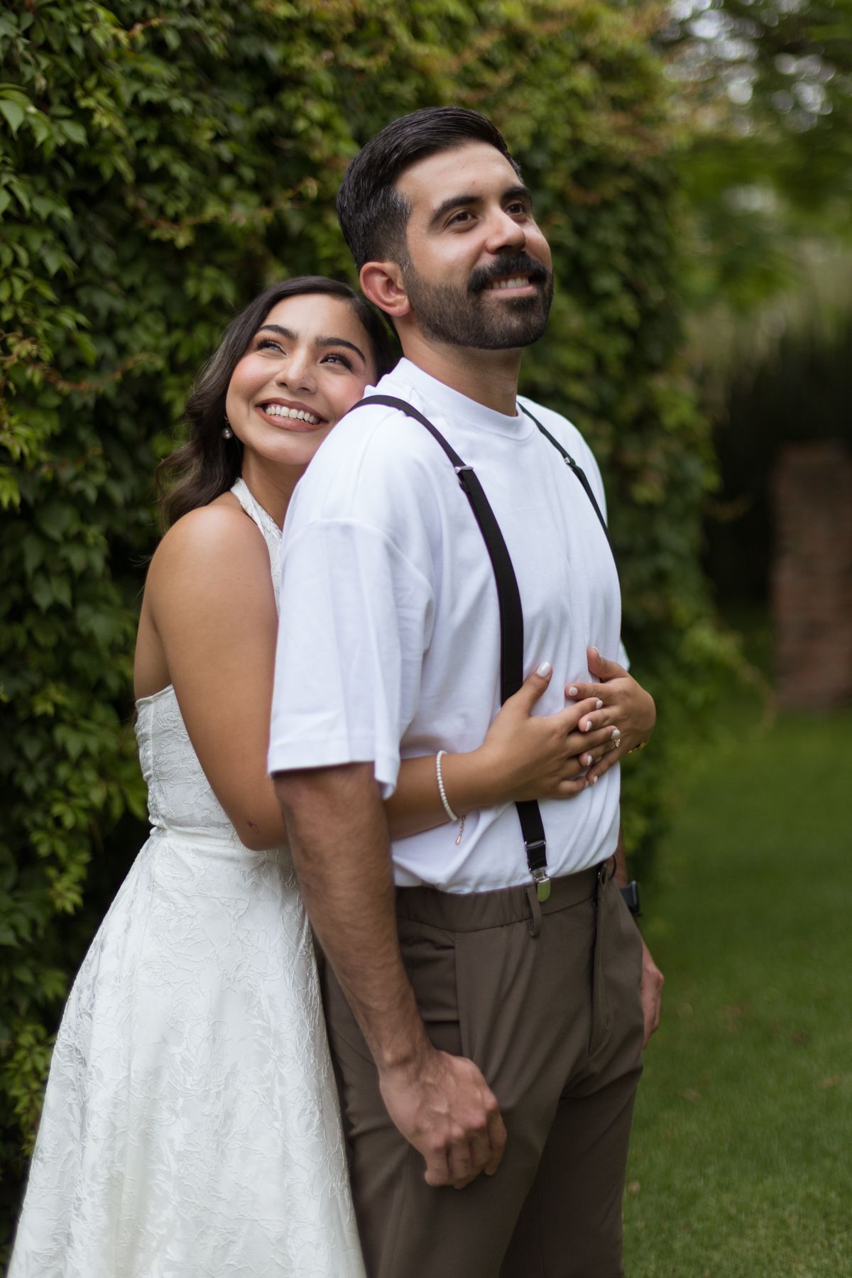 Smiling bride in white dress embraces groom in white shirt with suspenders in lush green garden.