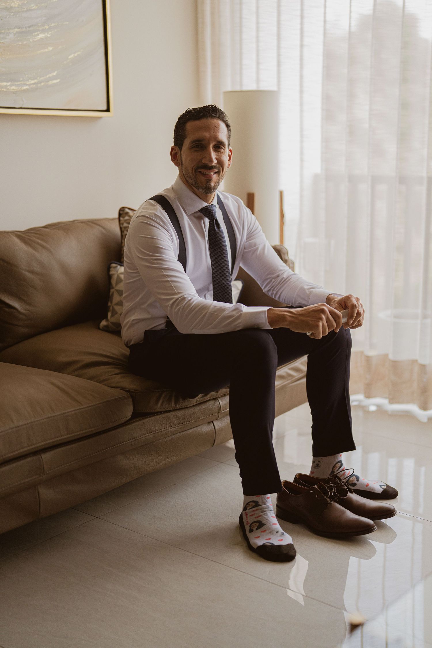 Smiling groom in suspenders and tie sits relaxed on leather sofa while getting ready for wedding.