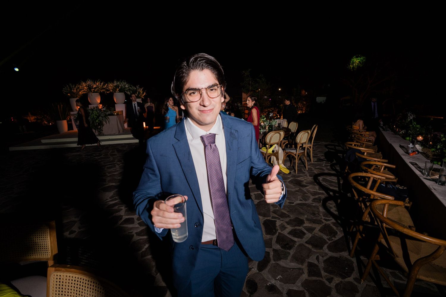 Smiling young man in blue suit gives thumbs up at outdoor wedding reception venue at night.