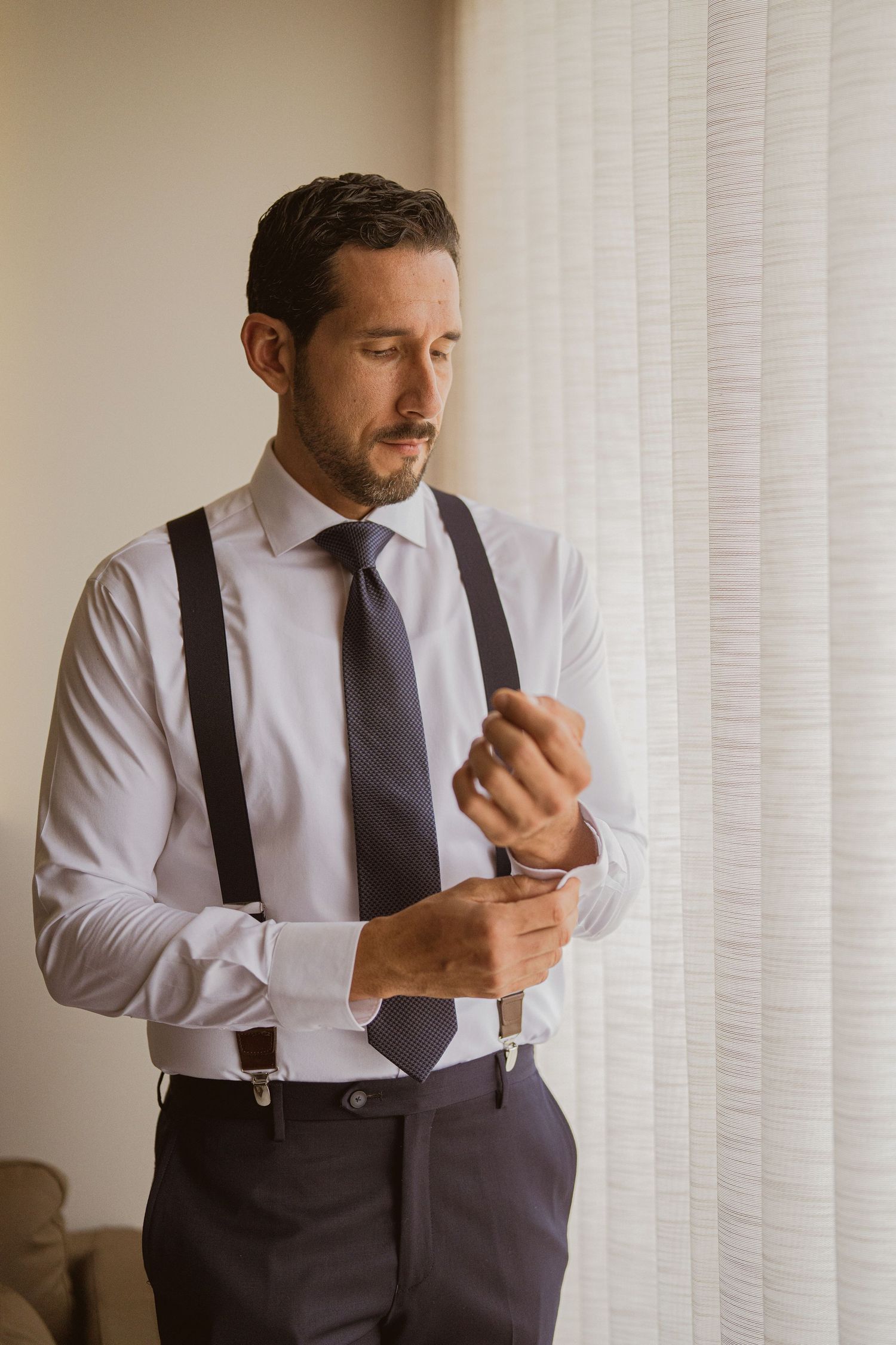 Groom in white shirt with suspenders fastening cufflinks by a bright window.