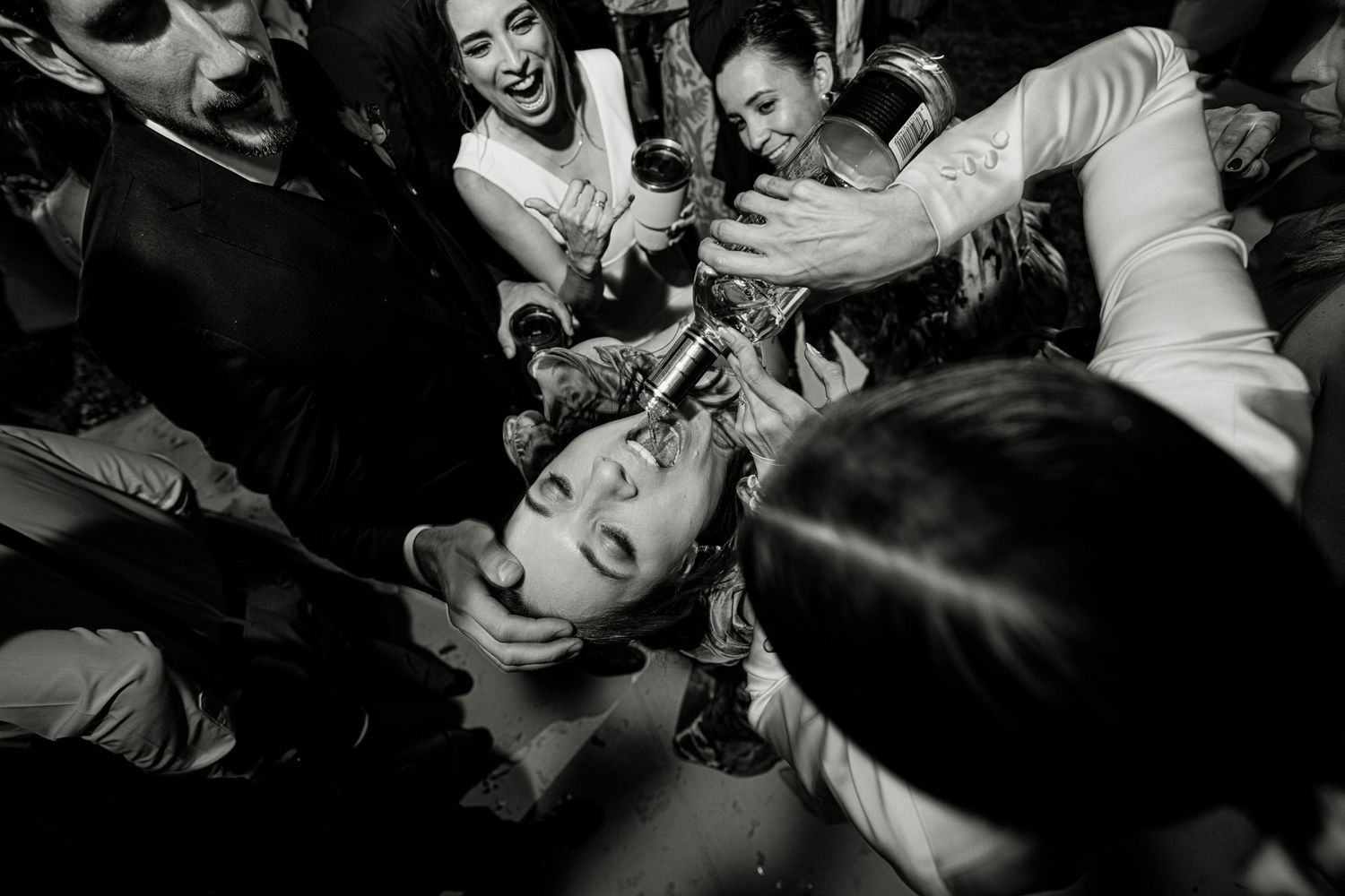 Wedding guests pour champagne into laughing persons mouth in fun black and white party photo.