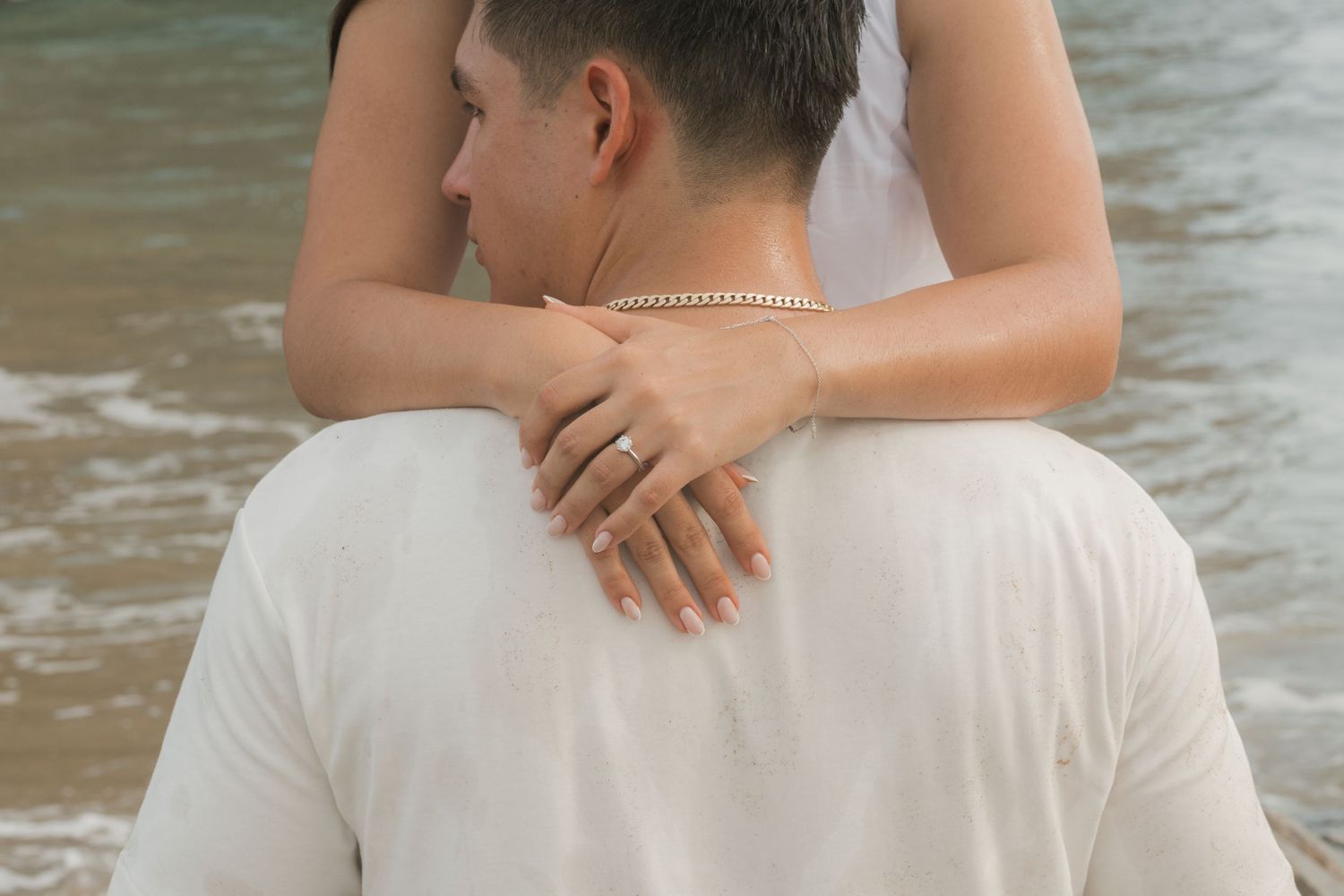 Couple embracing on a beach, woman wearing engagement ring resting hands on man's shoulders in white shirt.