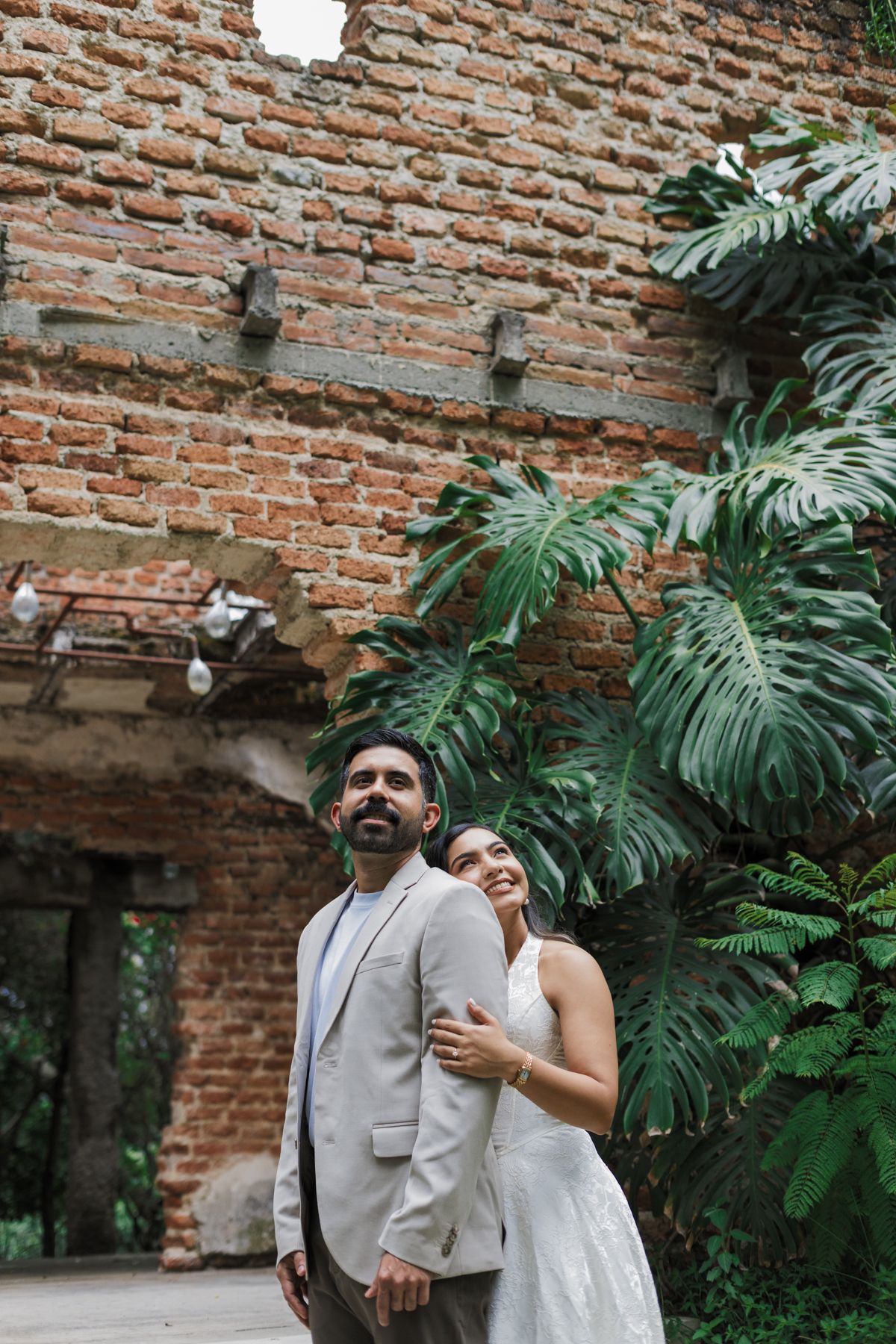 Couple in wedding attire stands before rustic brick wall with tropical monstera plants nearby.