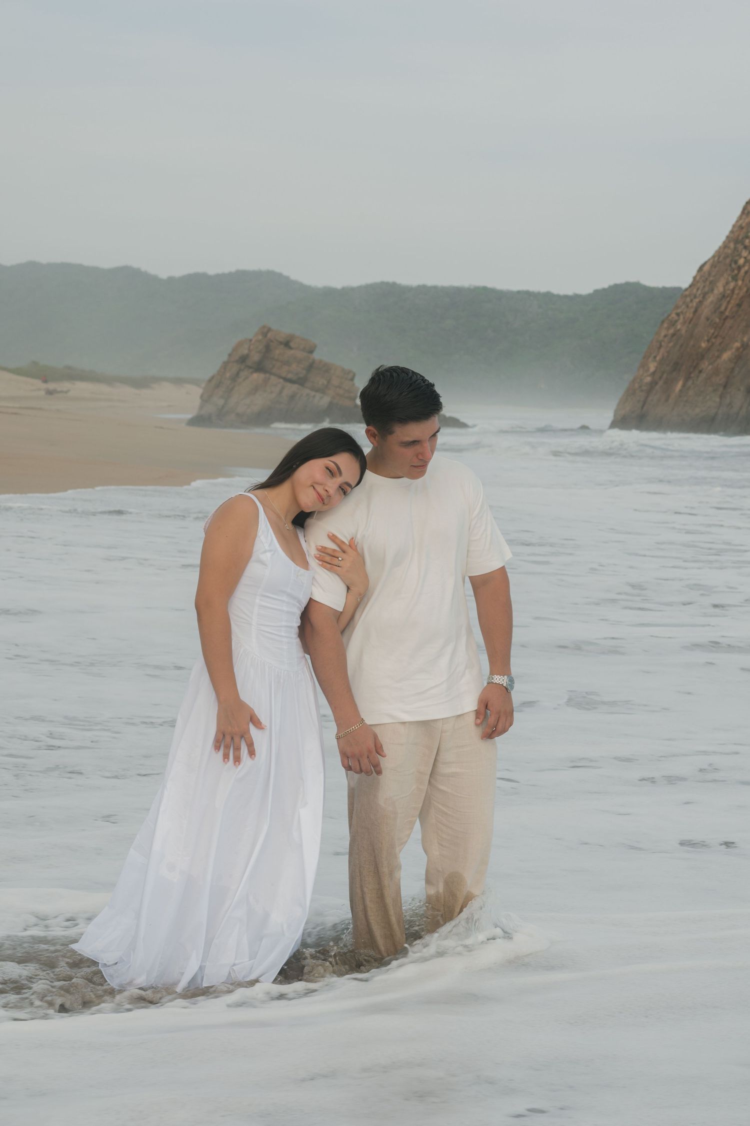 Romantic couple in white outfits walking along a misty beach shoreline with waves and rocky cliffs in the background.