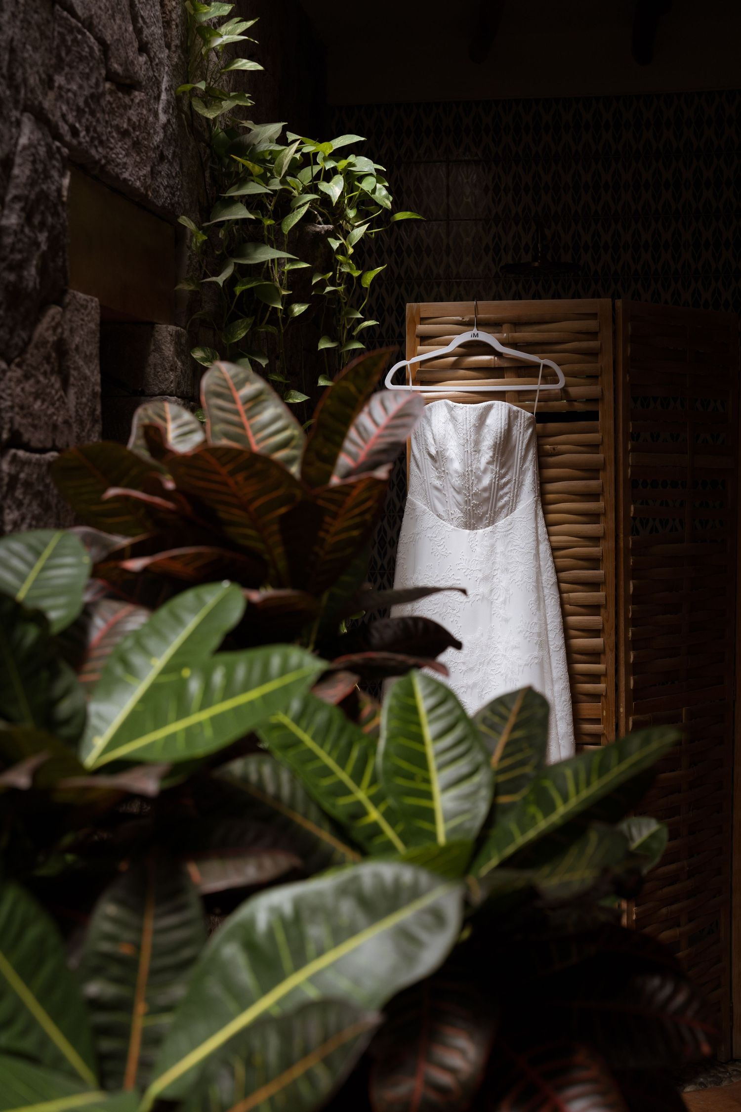 White strapless wedding dress hanging on wooden shutter surrounded by lush tropical green and red foliage.
