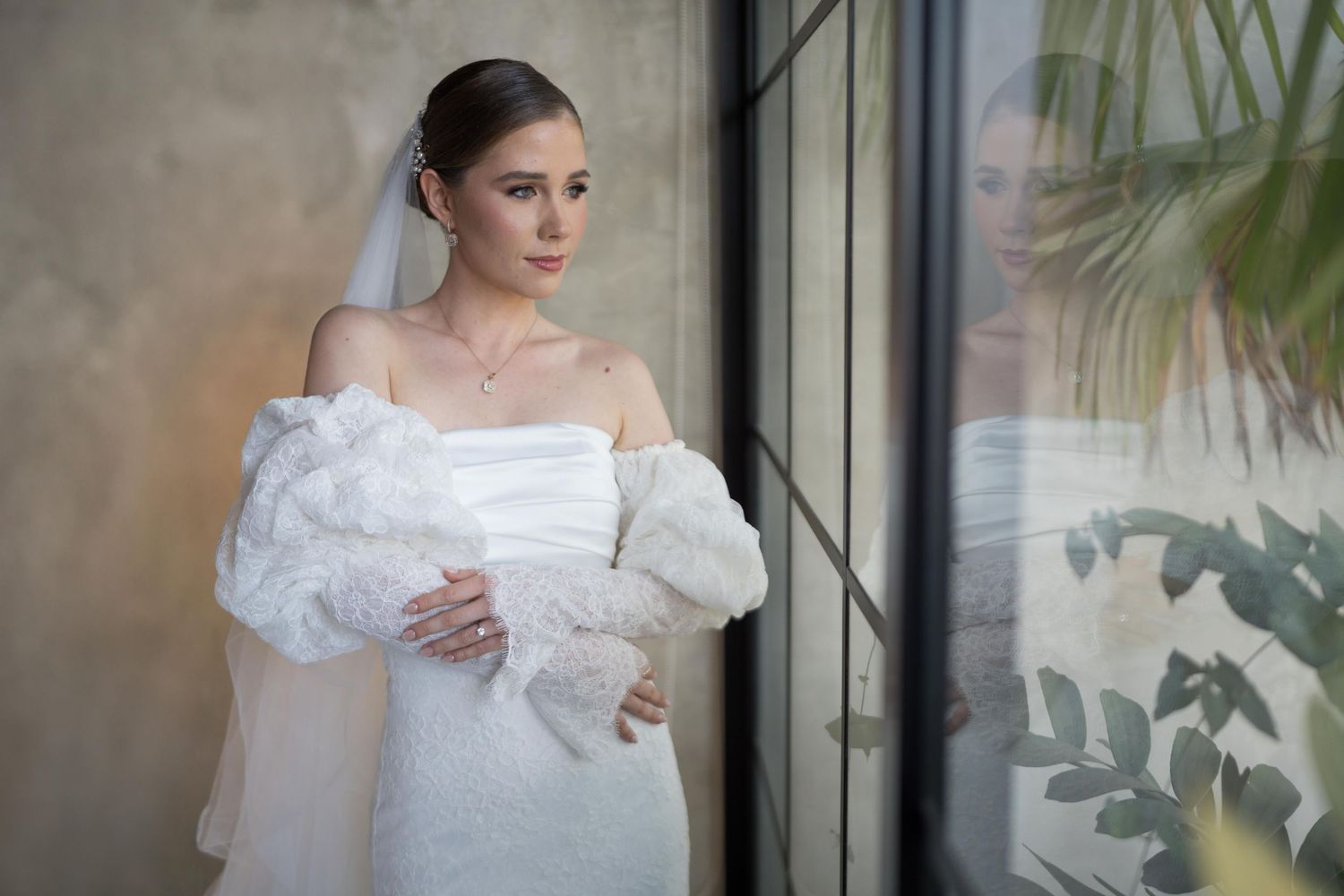 Beautiful bride in off-shoulder white gown with fluffy sleeves and veil gazes thoughtfully beside a sunlit window.