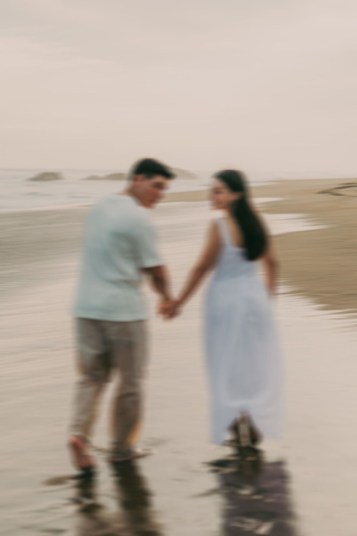 A couple holds hands walking barefoot on a sandy beach shore, the man in a light shirt and the woman in a white dress.
