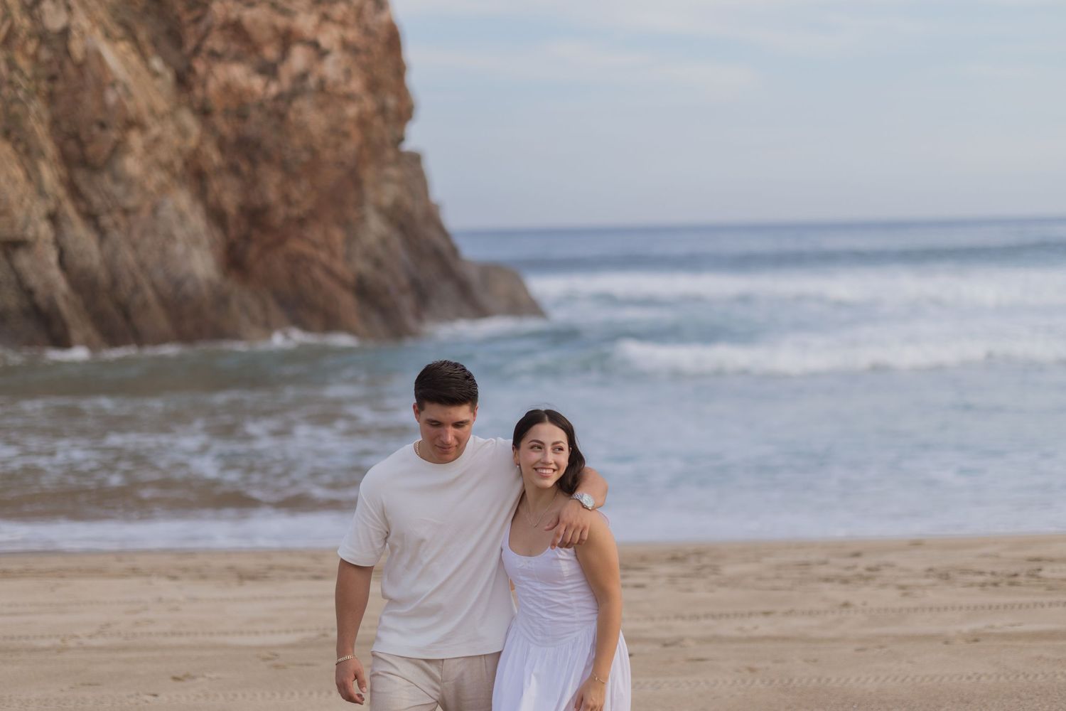 Couple walking on sandy beach with ocean waves and rocky cliffs, wearing white outfits in a romantic coastal setting.