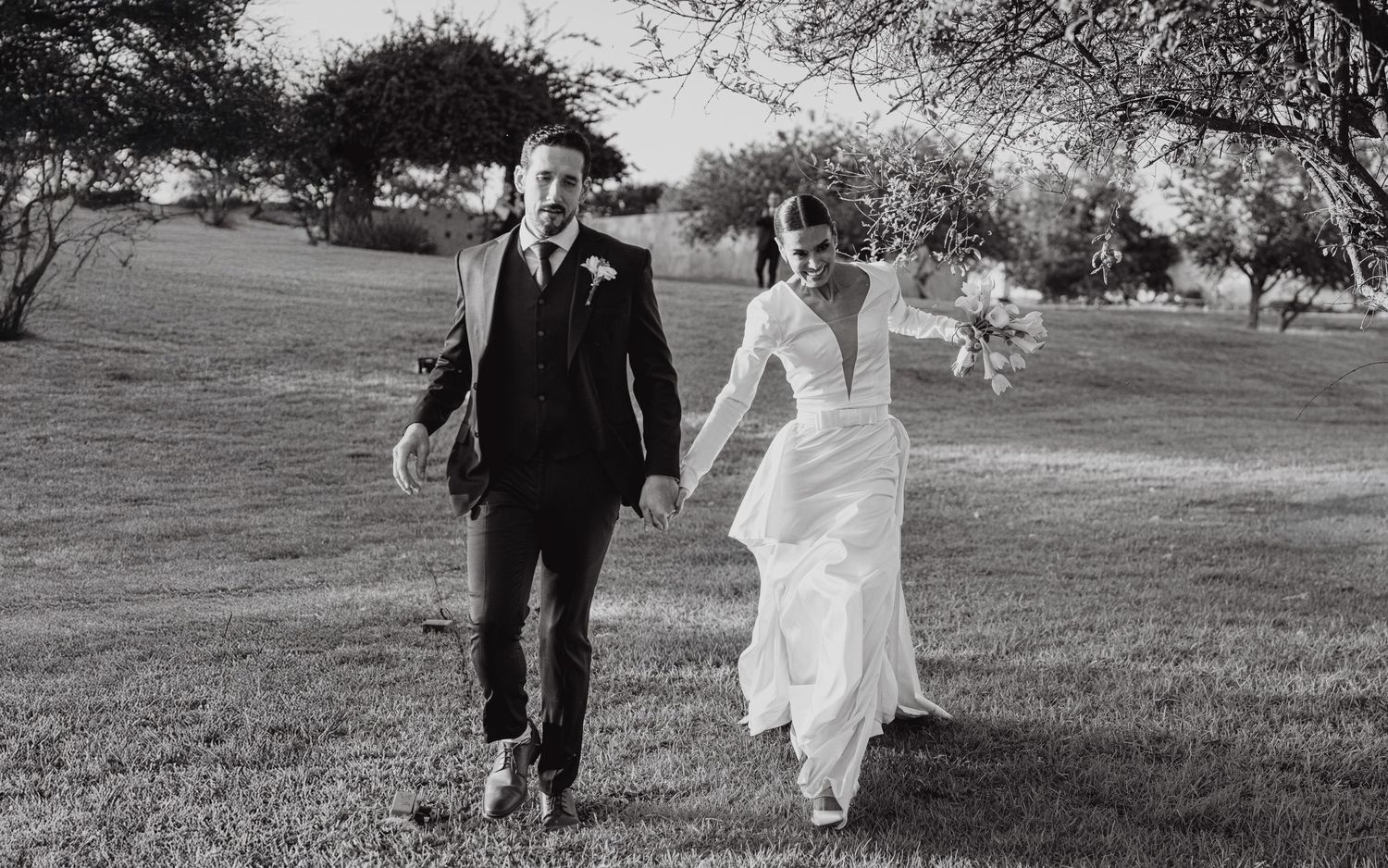 Black and white photo of bride and groom holding hands walking across green lawn.