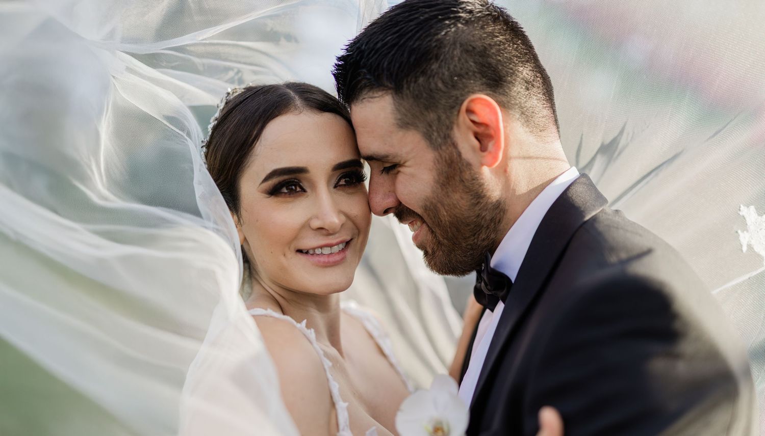 Stunning bride and bearded groom share a tender close moment framed by a billowing white veil outdoors.