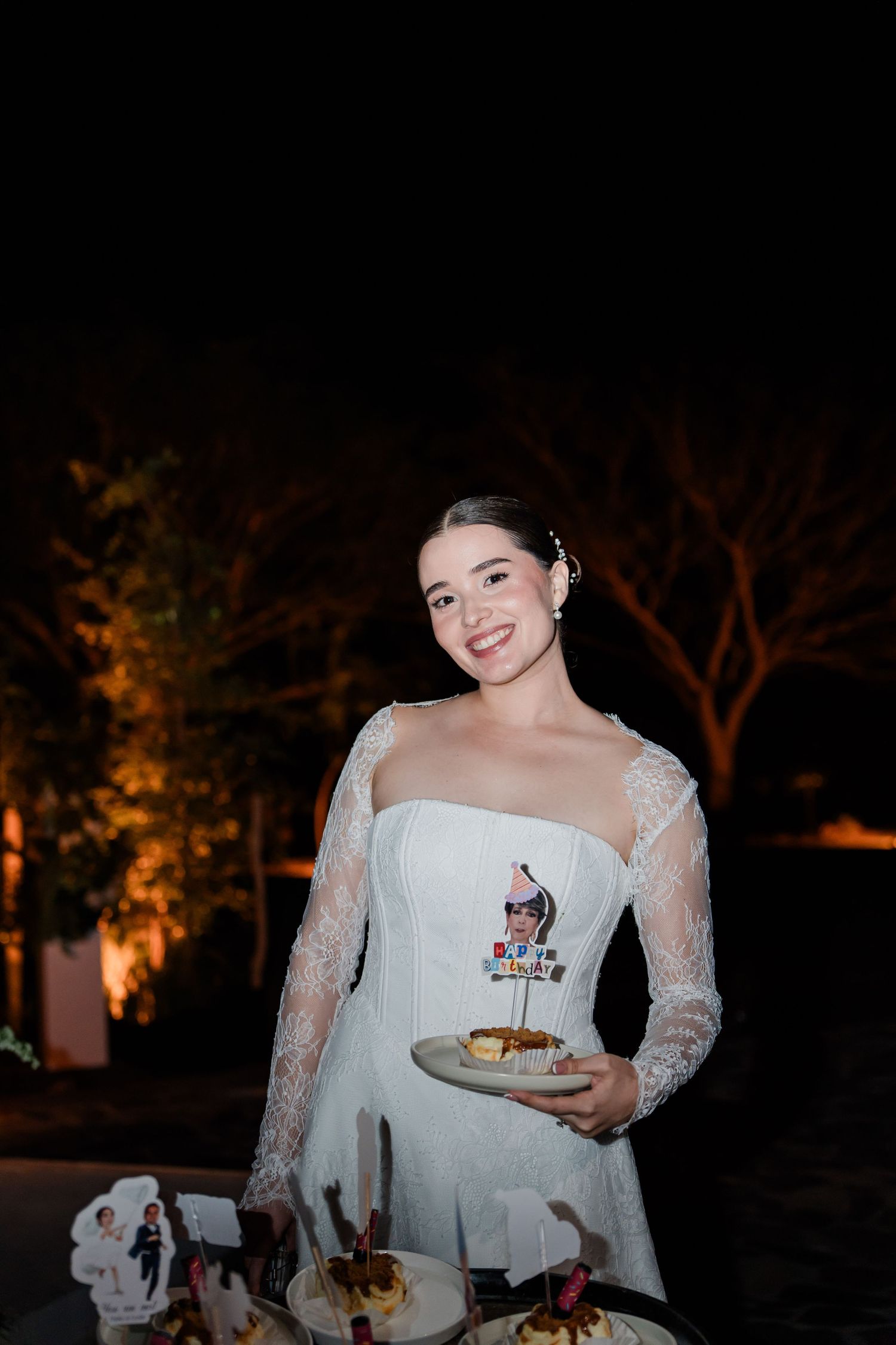 Smiling bride in lace wedding gown holding dessert plate at outdoor nighttime wedding reception.
