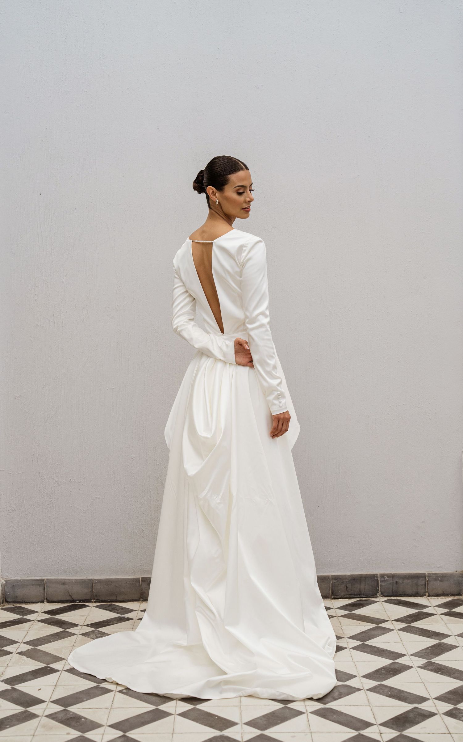 Bride in long-sleeve open-back white satin wedding dress standing on checkered tile floor.