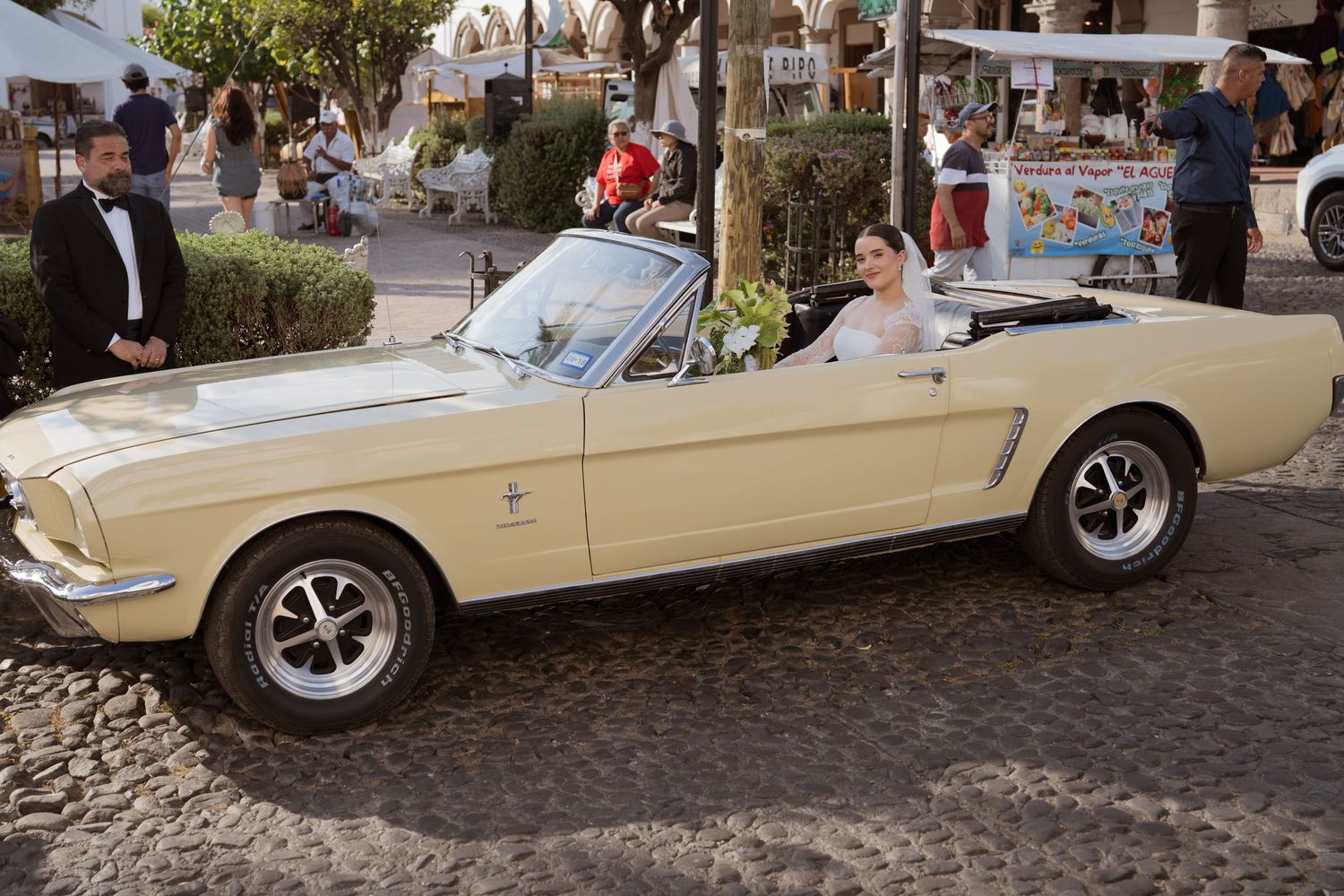 Radiant bride arrives in a classic cream vintage Ford Mustang convertible on a sunny wedding day.