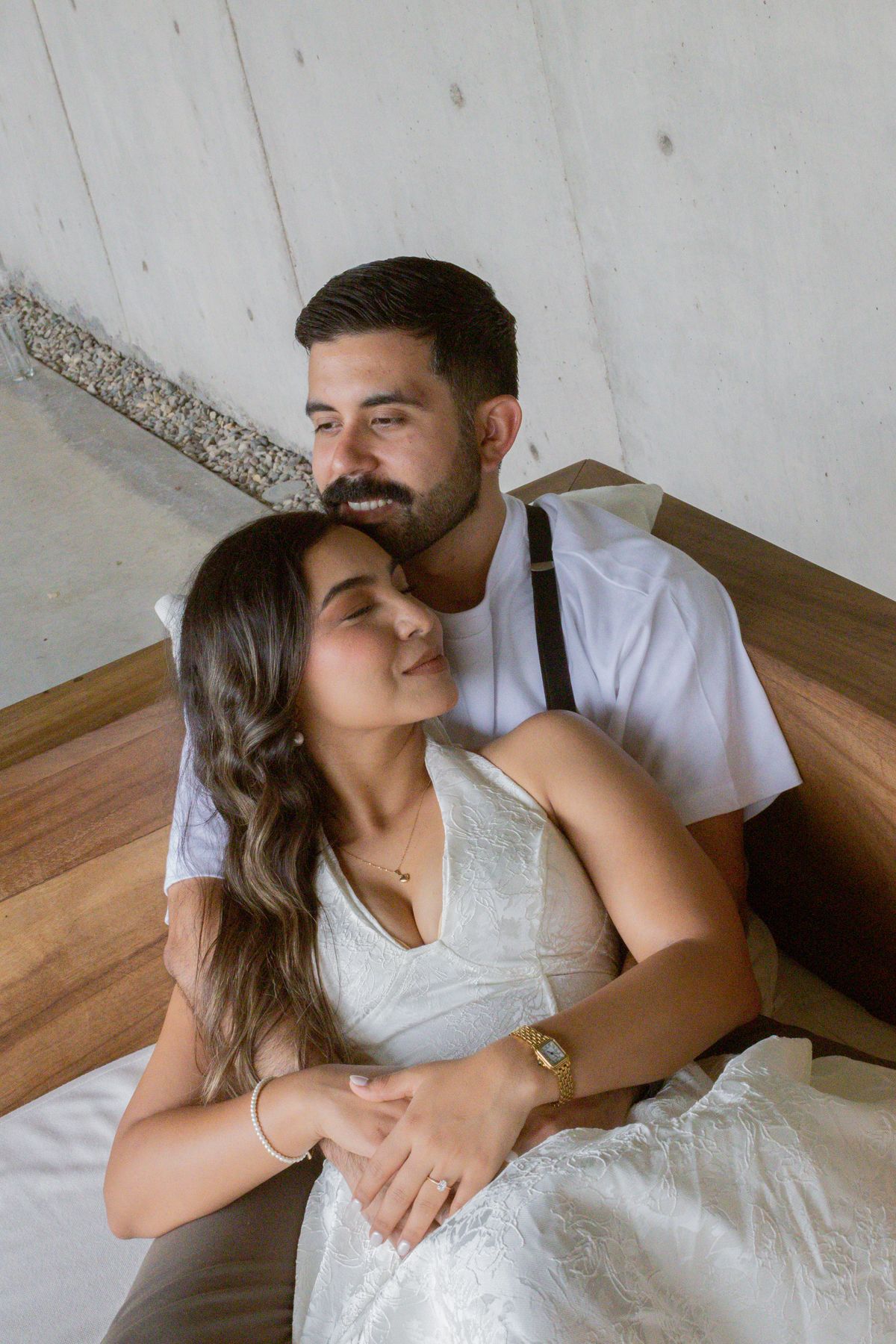 Couple embracing on a bed, woman in white dress leaning back against smiling bearded man in white tee.