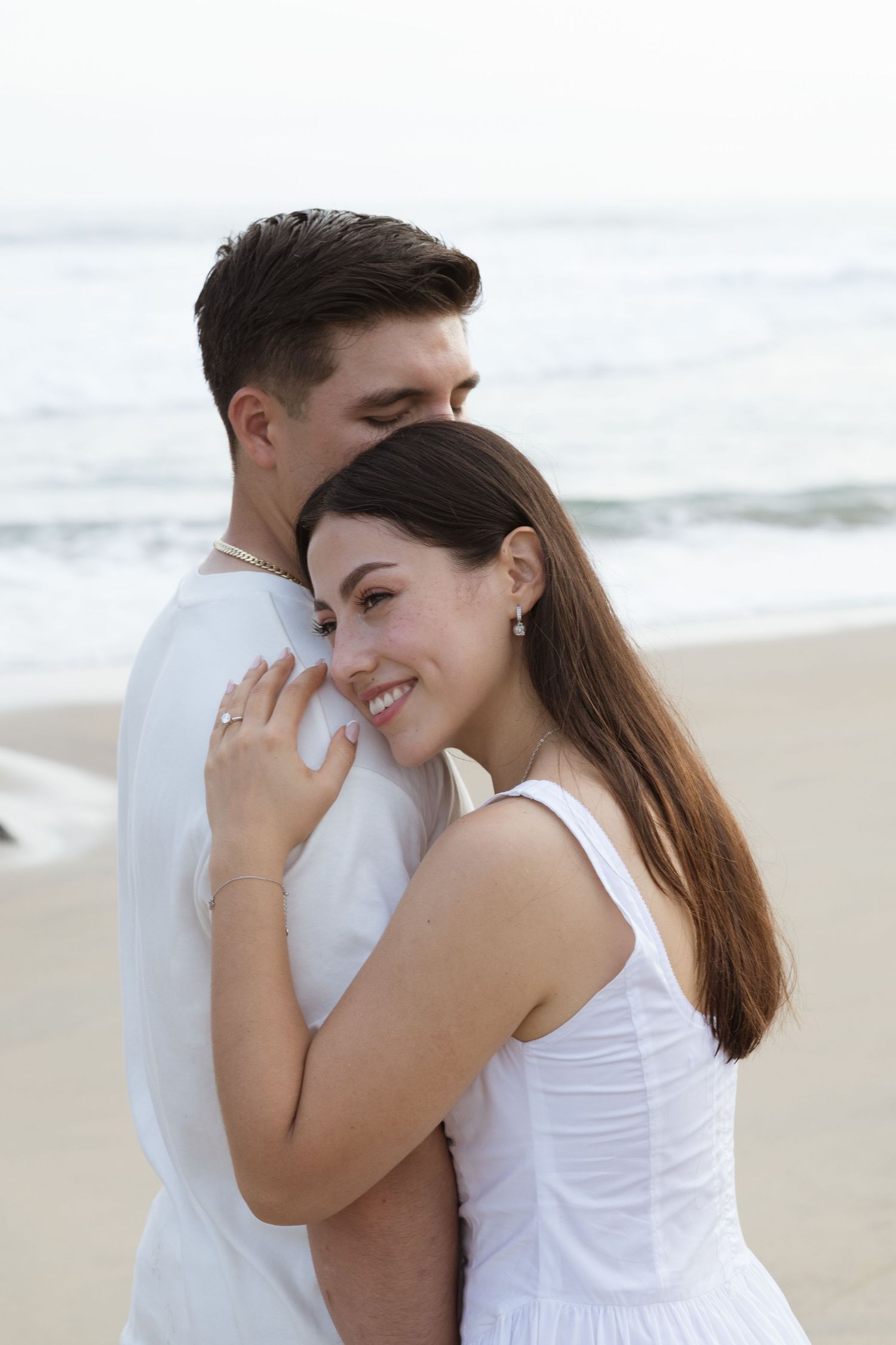 A smiling couple in white outfits embracing on a sandy beach with ocean waves in the background.