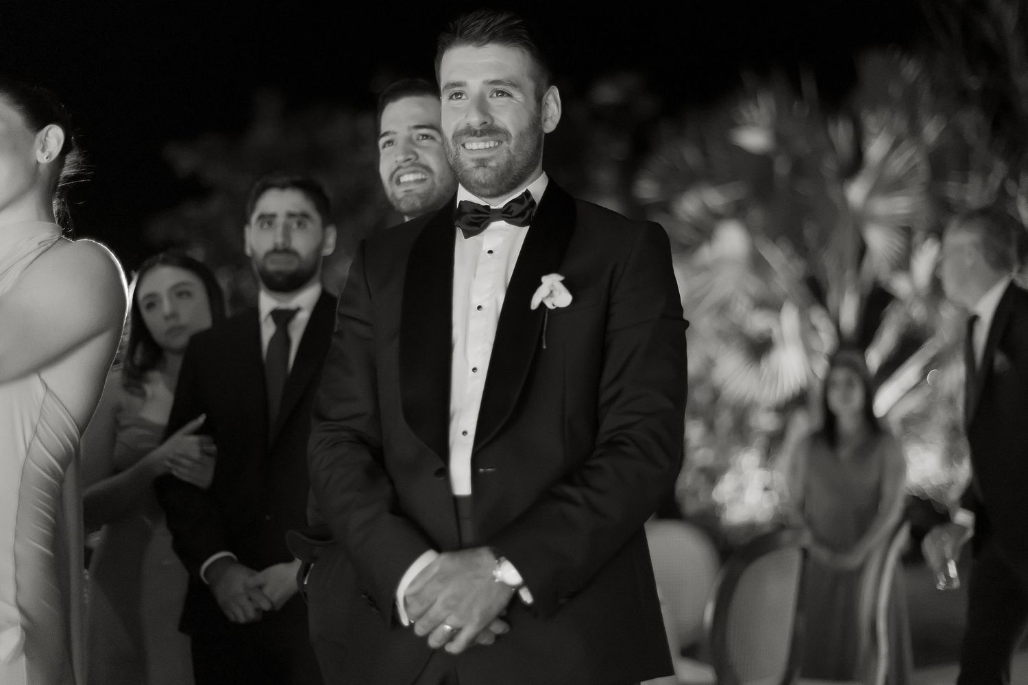 Groom in black tuxedo smiling with clasped hands at wedding ceremony altar in black and white photo.