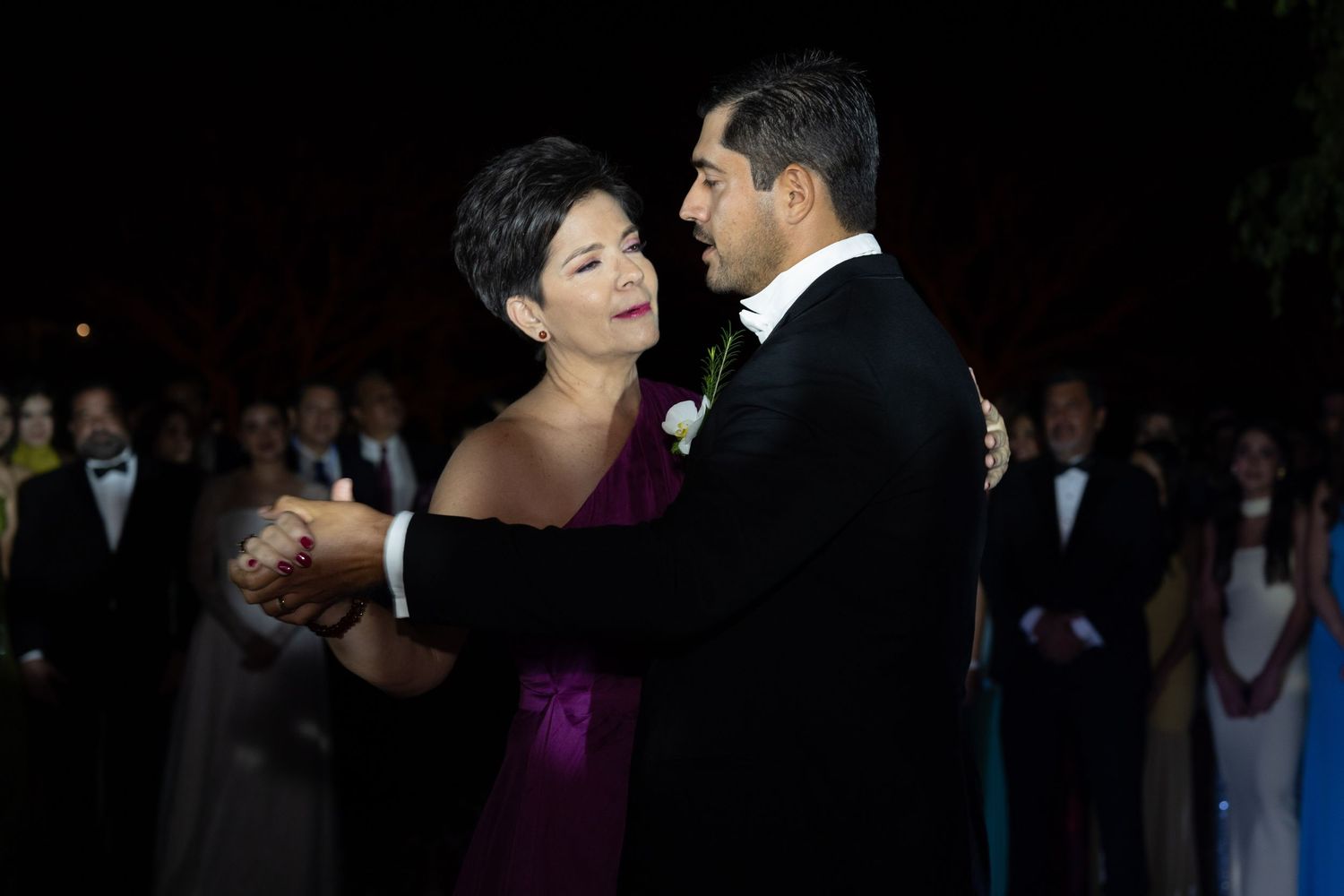 Groom shares a tender mother-son dance at an elegant outdoor wedding reception with guests watching.