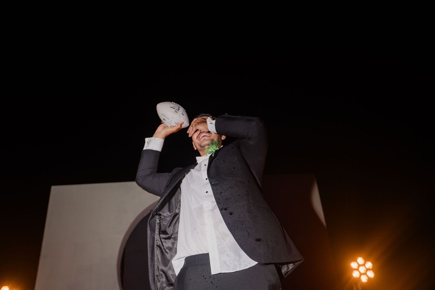 Groom in black suit playfully holds a white object above his head under bright stage lights at a wedding reception.
