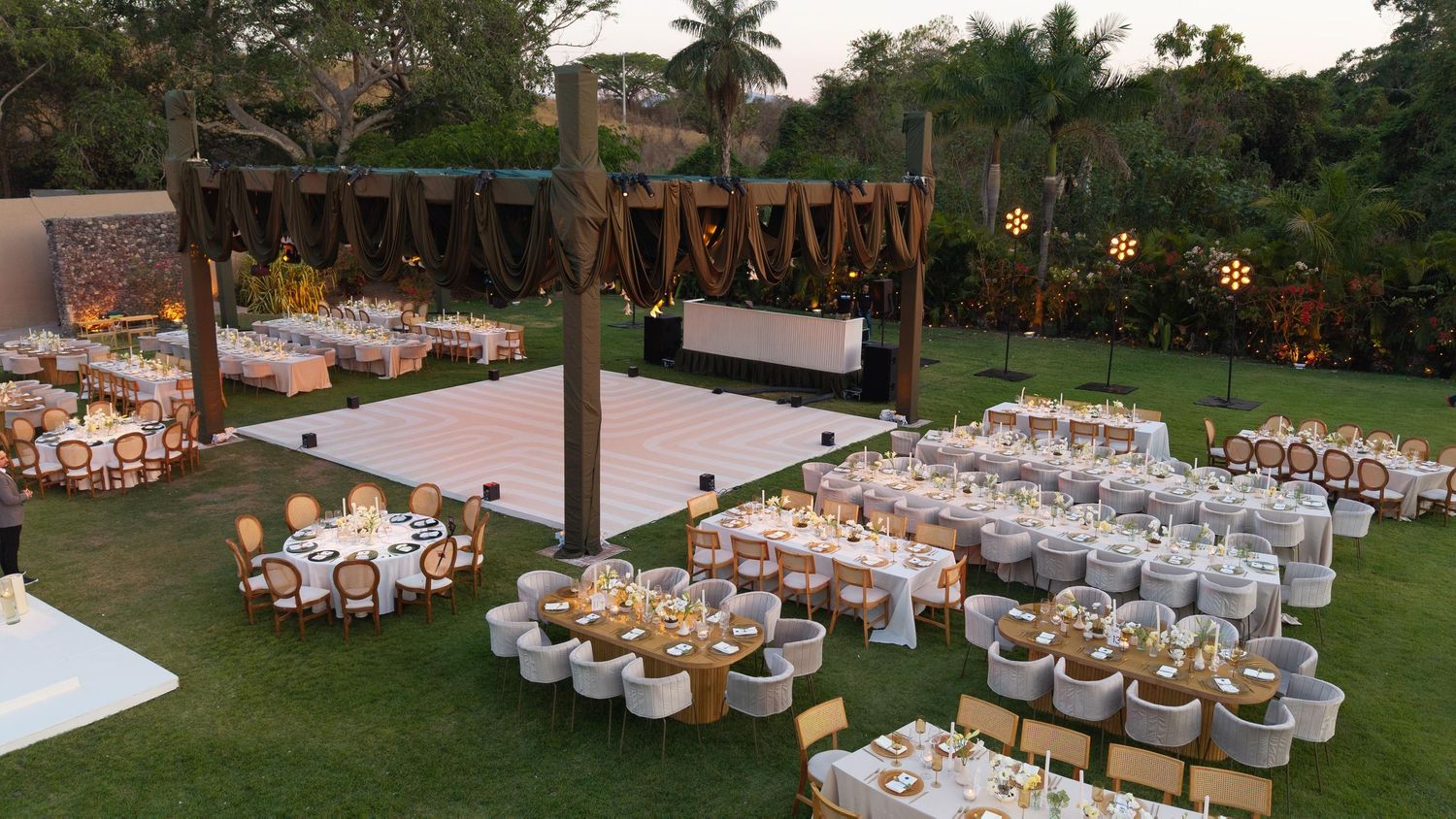 Bird's-eye view of a lush garden wedding venue featuring round tables, a dance floor, and rustic wooden pergola.