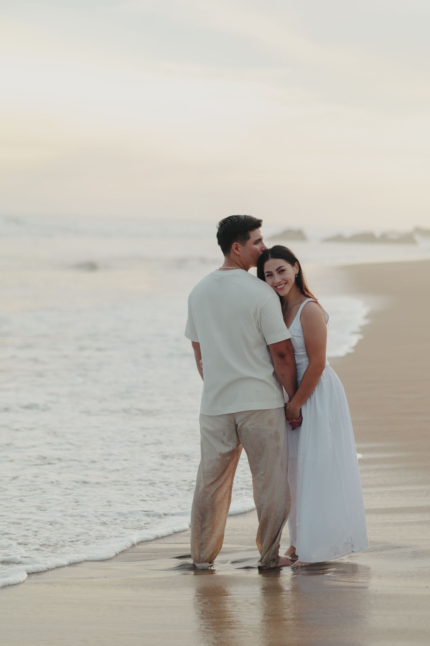 Romantic couple holding hands on a sandy beach at sunset, woman in white dress smiling, man in light casual outfit.