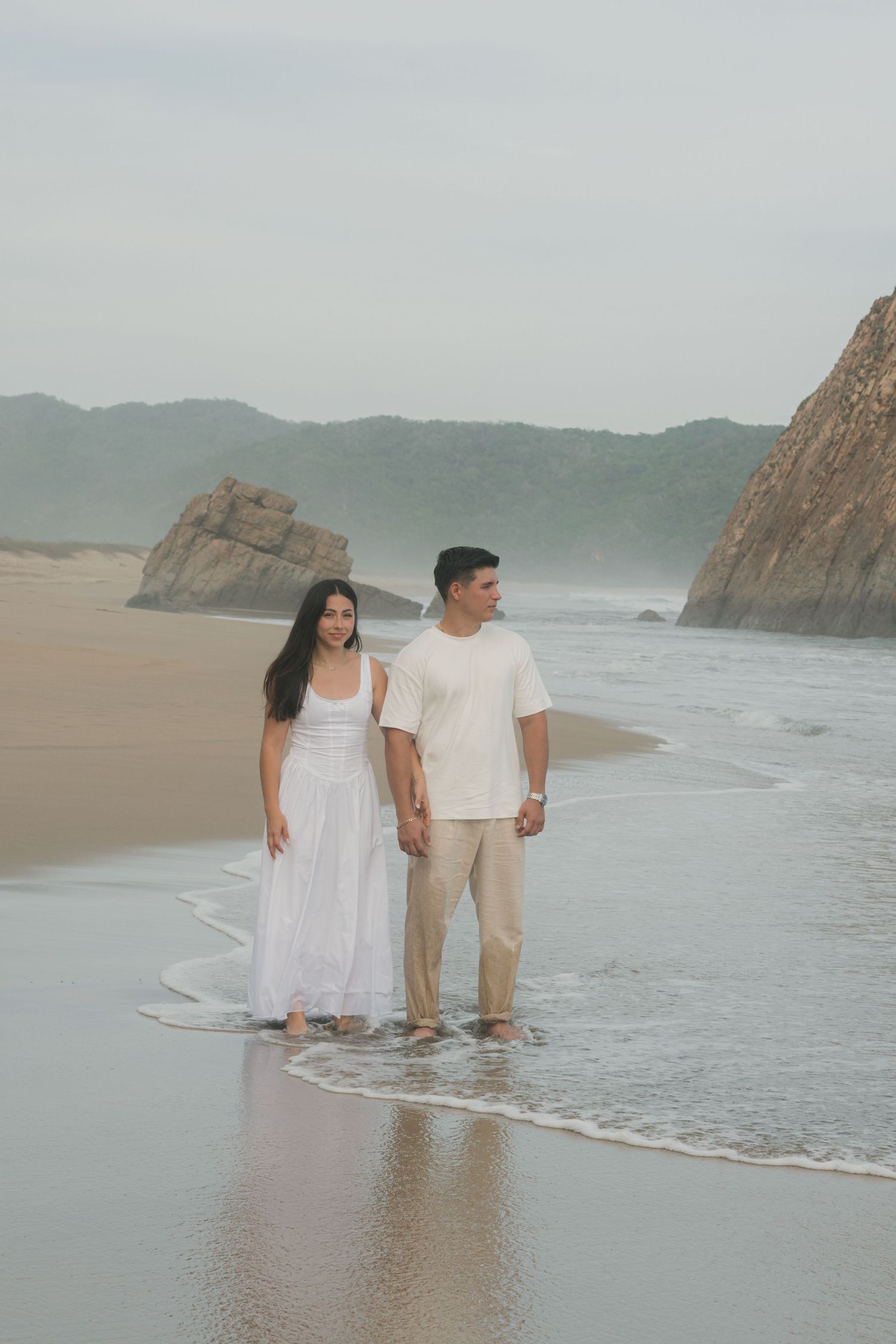 Couple holding hands on a misty beach, woman in white dress, man in cream outfit, with rocky cliffs in the background.