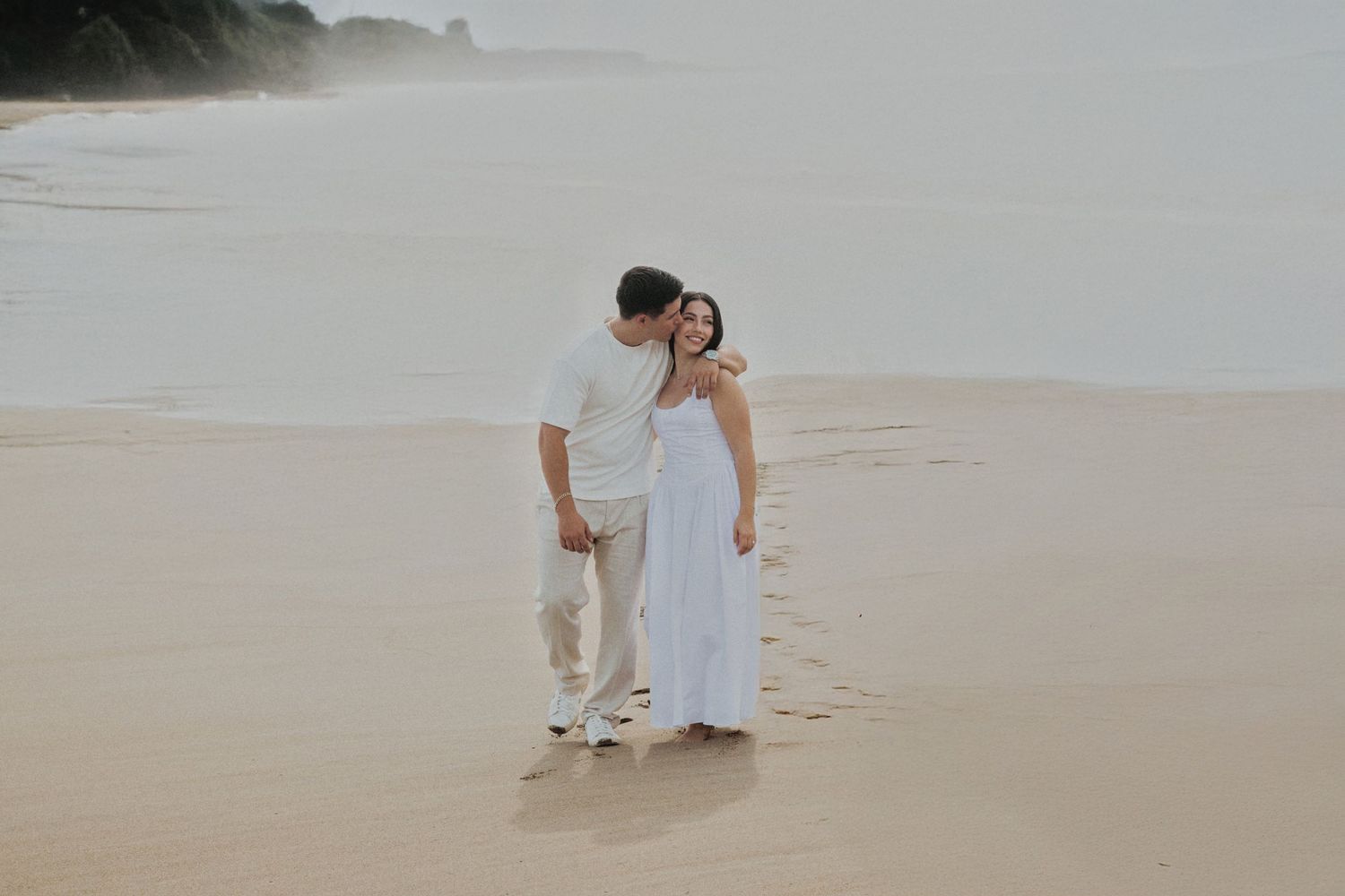 Couple in white attire walking hand in hand on a sandy beach with misty ocean waves in the background.