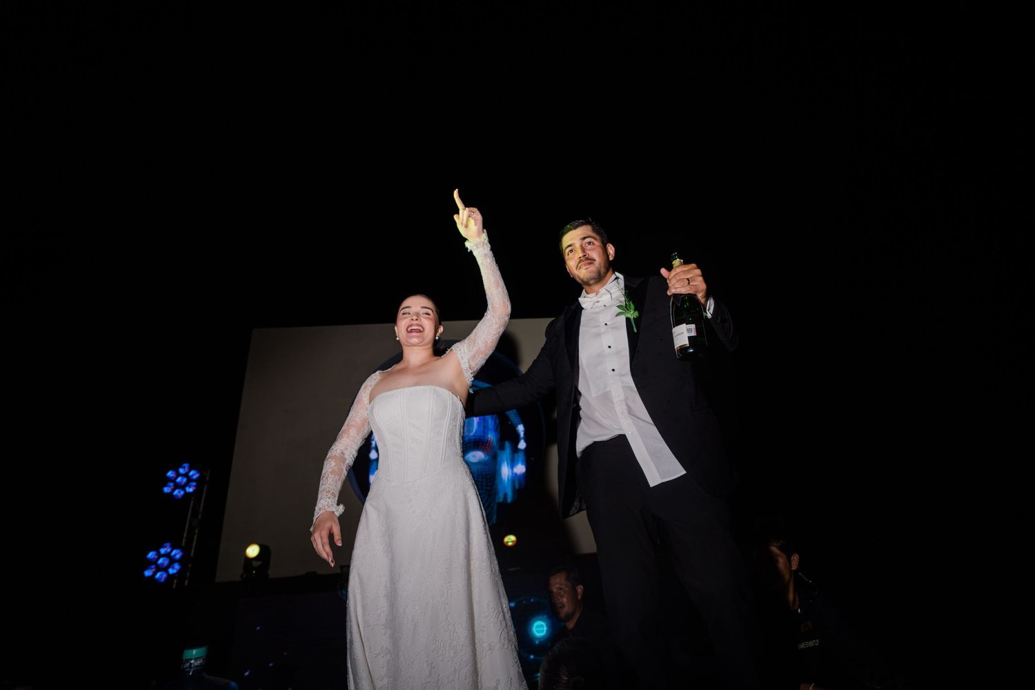 Bride and groom celebrate joyfully on stage at night, raising their hands with sparklers at their wedding reception.