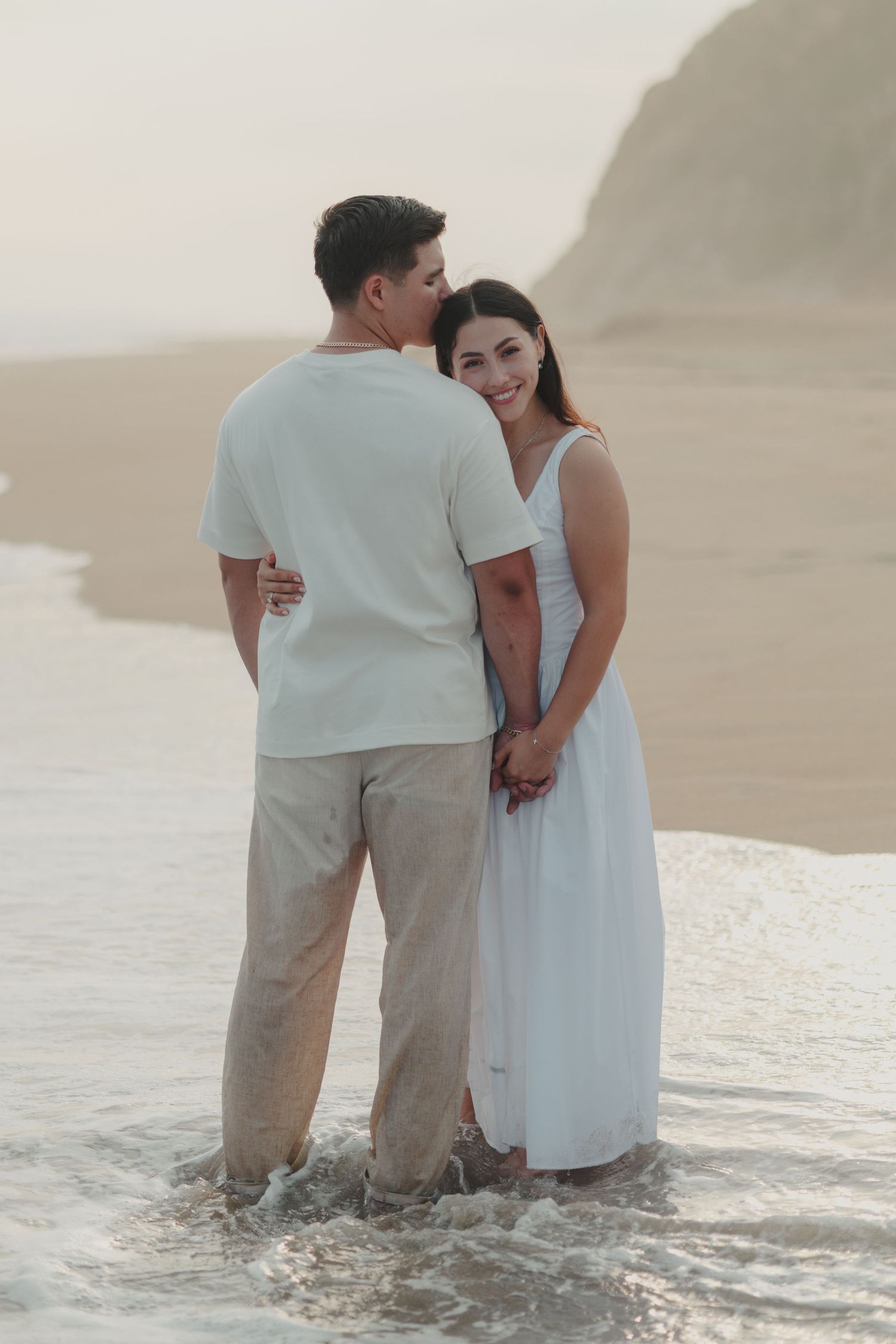 Couple standing in ocean waves at beach, woman in white dress smiling, man in white shirt and tan pants holding hands at sunset.