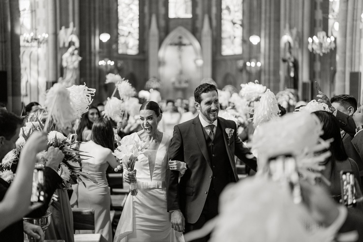 Black and white photo of a joyful couple walking down a church aisle surrounded by feathered bouquets.