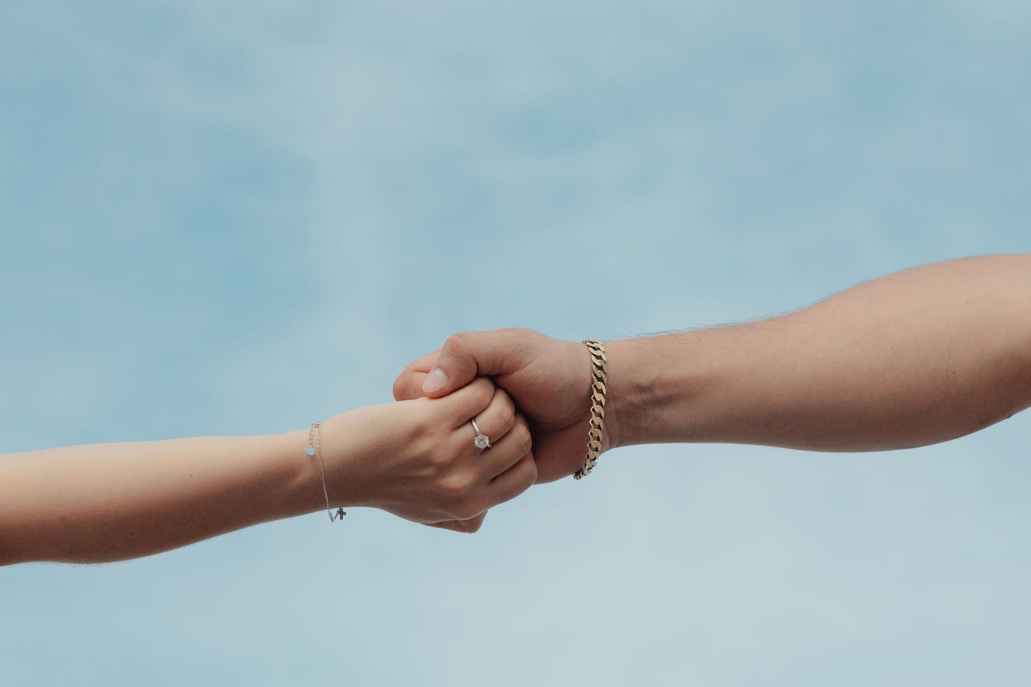 Two hands clasping together against a blue sky, one wearing a gold chain bracelet and a ring.