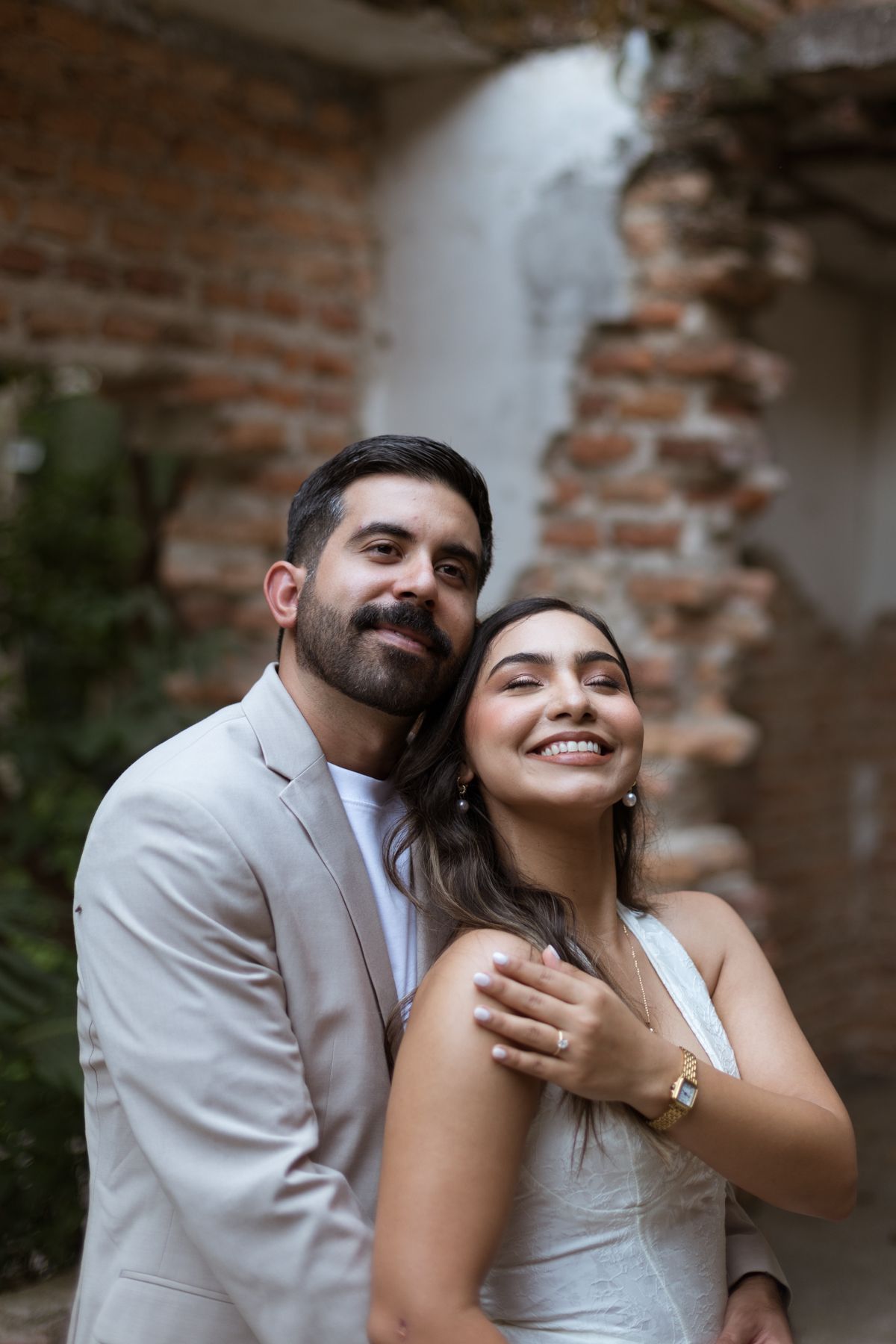 Happy couple embraces near rustic brick wall, bride in white dress with engagement ring visible.