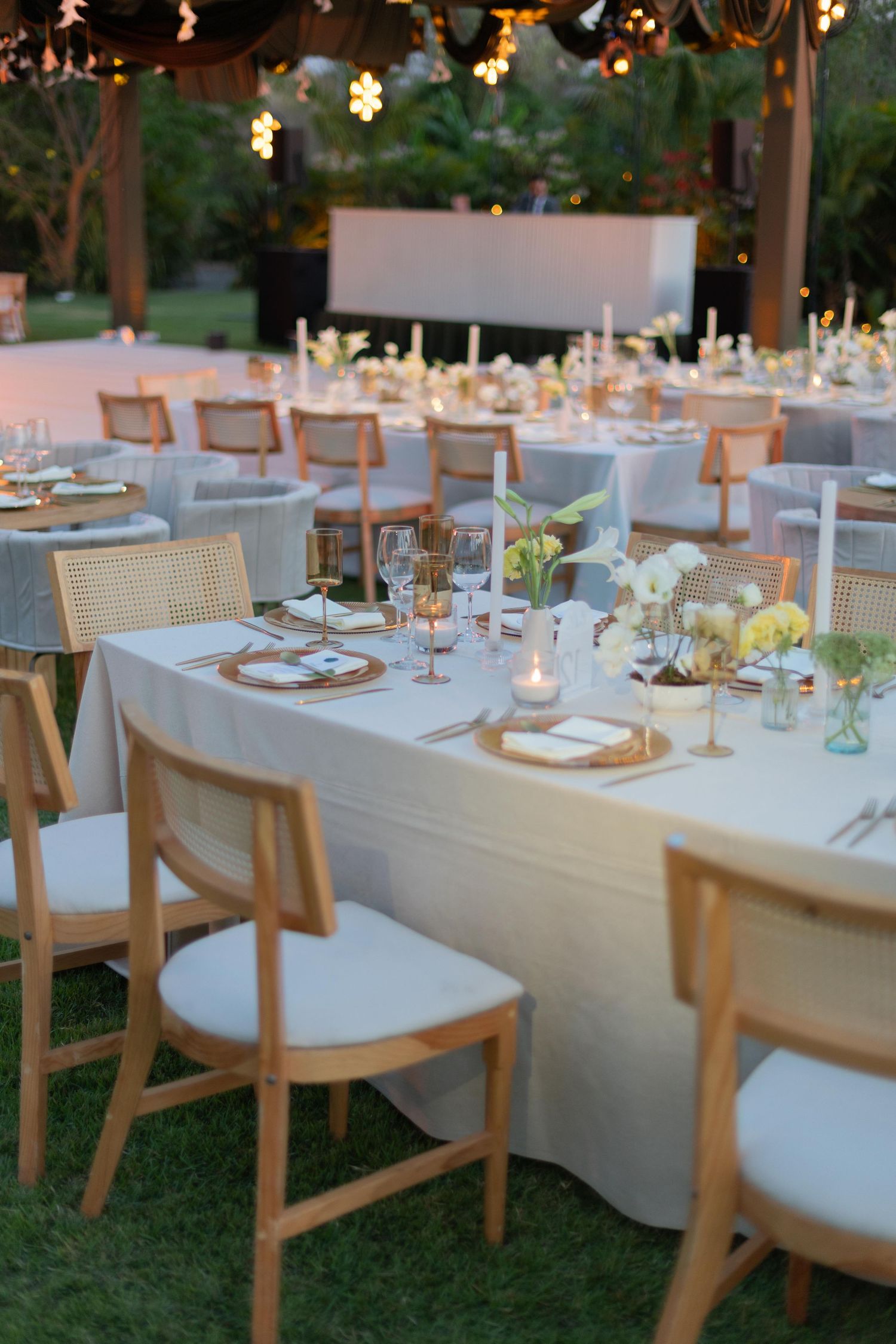 Elegant outdoor wedding reception with wooden chairs, white linen tables, candles, and floral centerpieces at dusk.