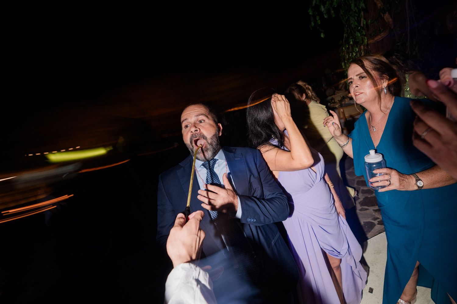 Man playfully biting a stick at an outdoor nighttime wedding celebration with guests.