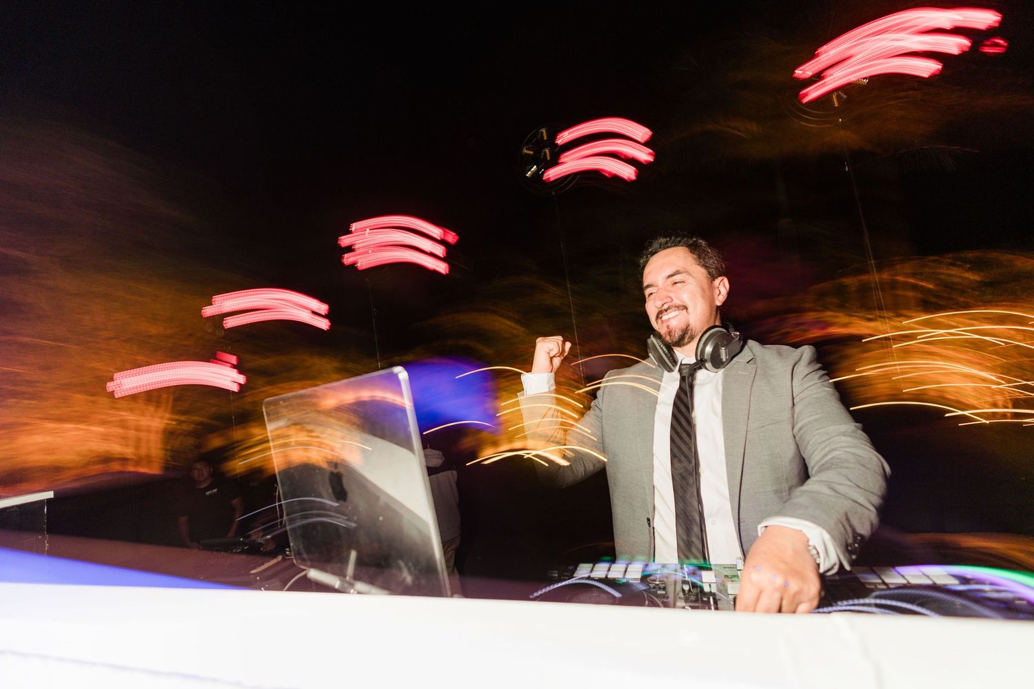 Smiling DJ in a suit performing at a wedding reception with dynamic light trails and colorful bokeh effects.