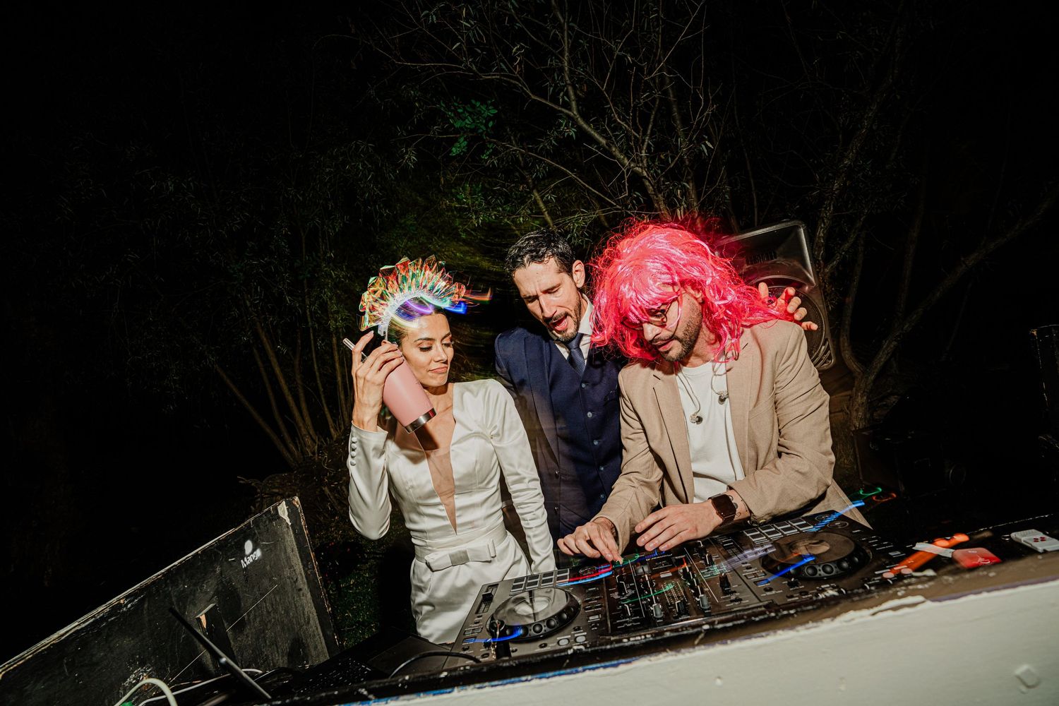 Bride in white gown with decorative headpiece joins DJ booth with groom at wedding after-party.