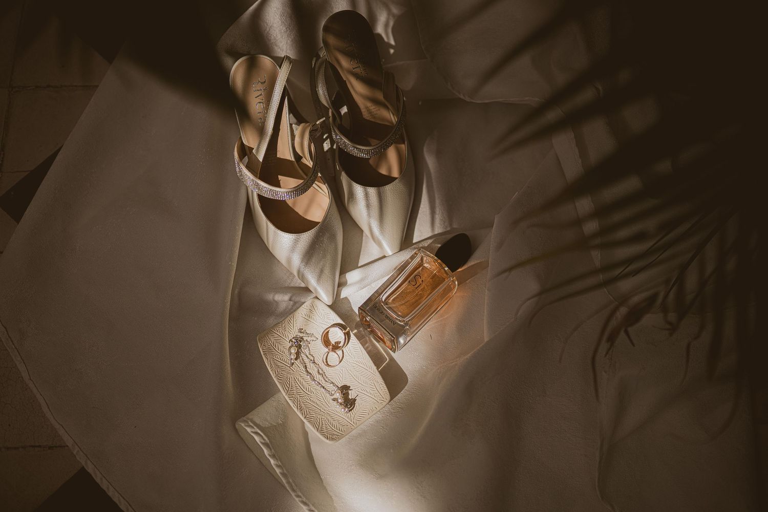 Elegant bridal flat lay with gold sandals, perfume bottle, and jewelry on white fabric.