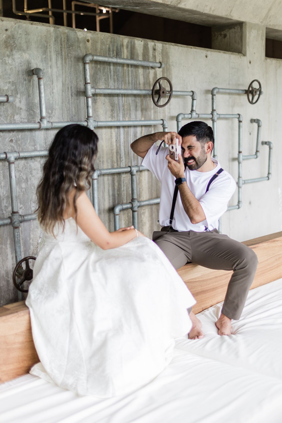 Groom photographing bride with a camera on a bed in an industrial-style room with exposed pipes.