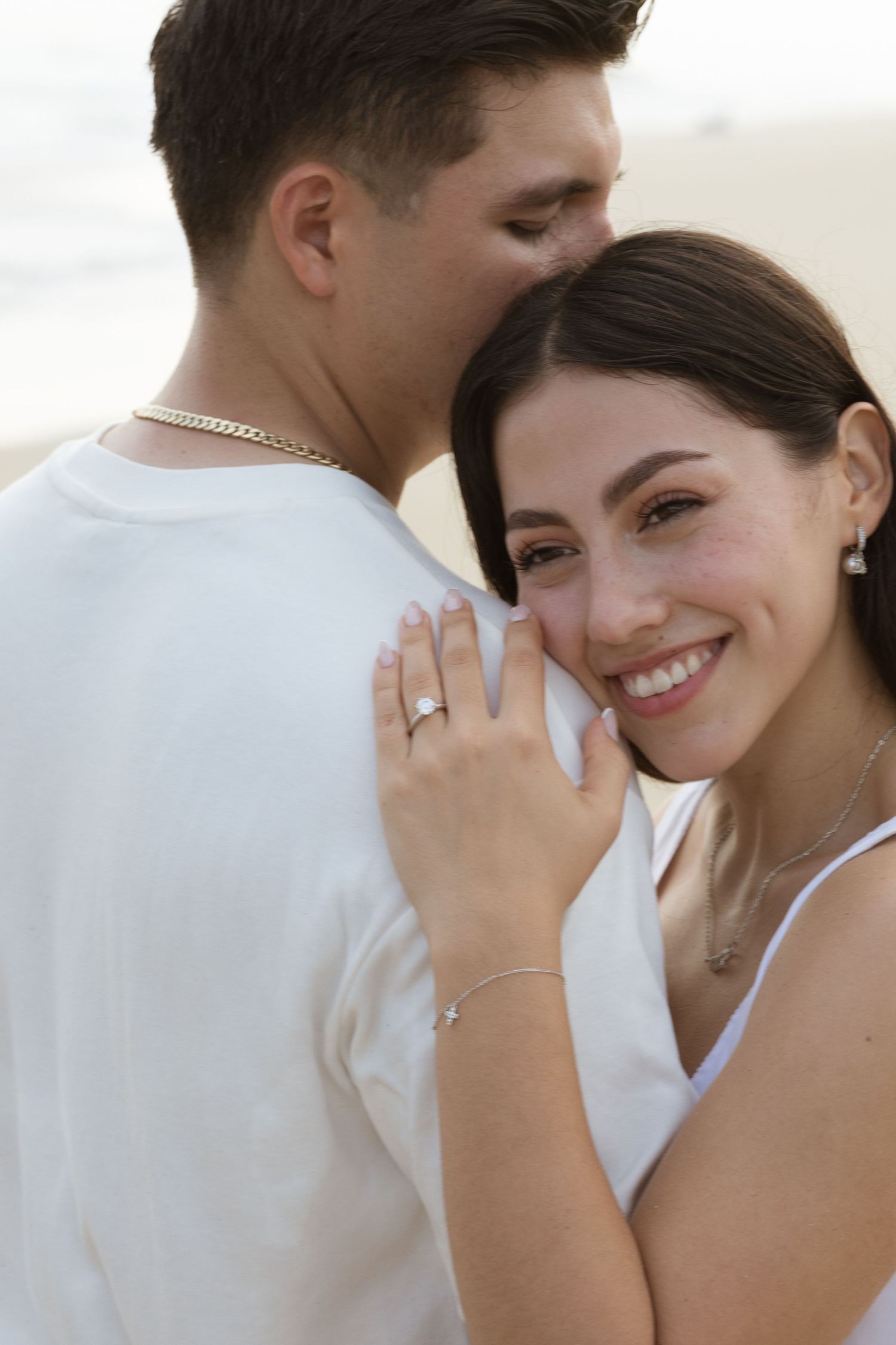 A smiling woman with an engagement ring embraces a man kissing her cheek on a beach.