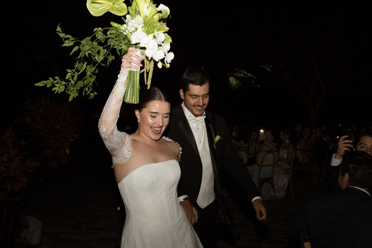 Smiling bride lifts her white floral bouquet as she walks with groom at their outdoor nighttime wedding.