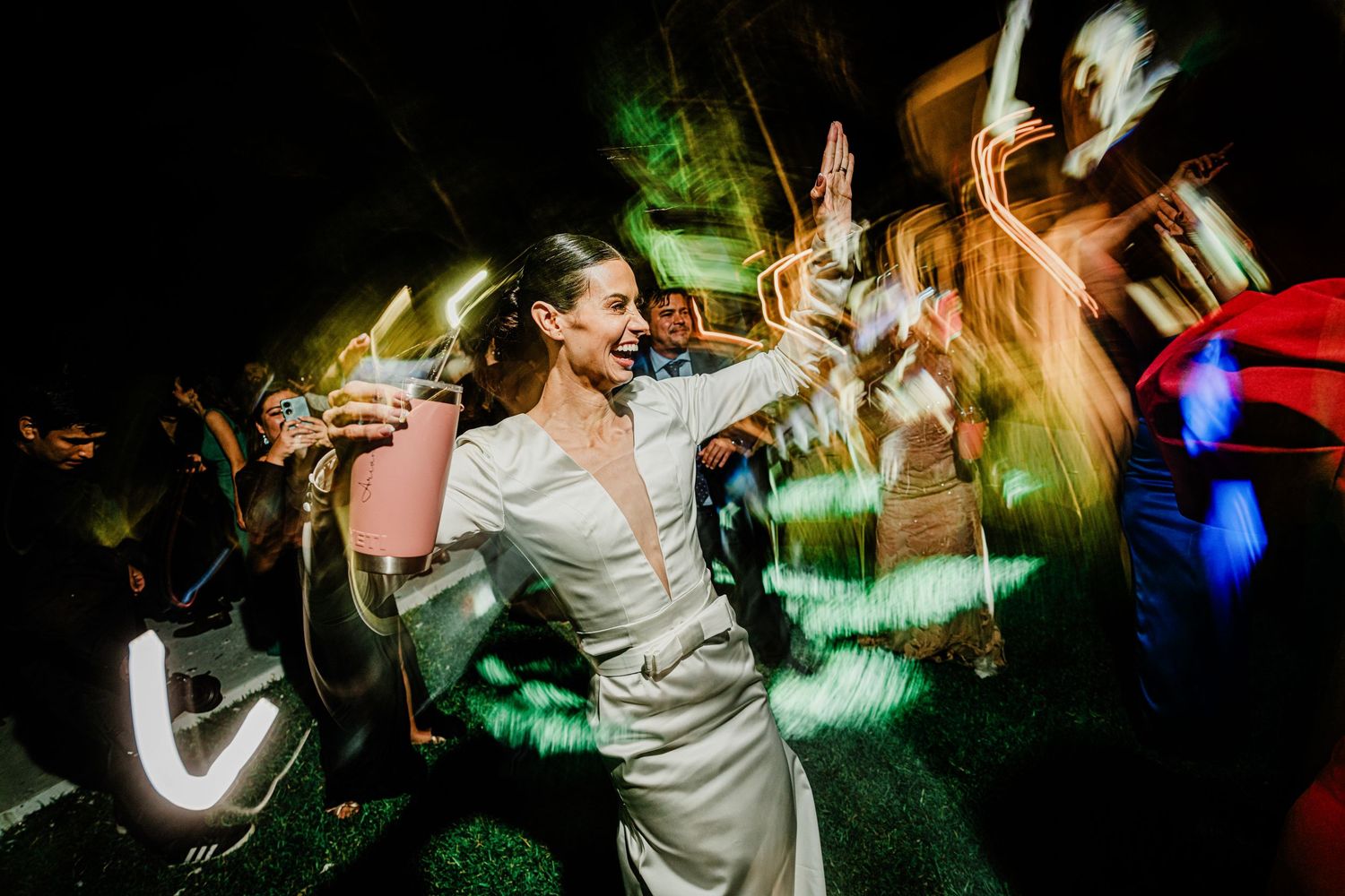 Smiling bride in white dress dancing with drink at vibrant outdoor wedding reception at night.