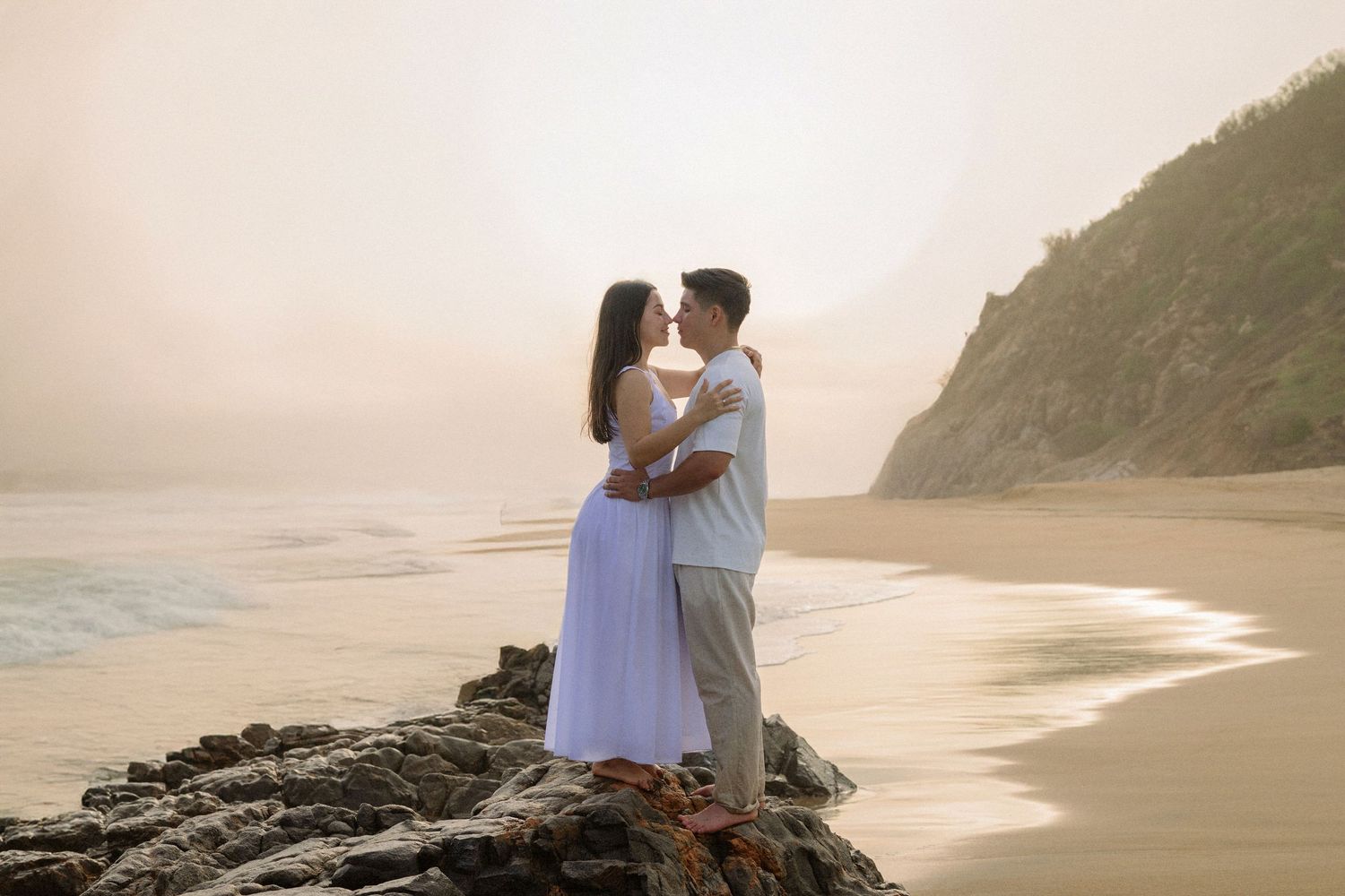 Couple embracing and kissing on rocky beach at sunset with ocean waves and misty cliffside backdrop.