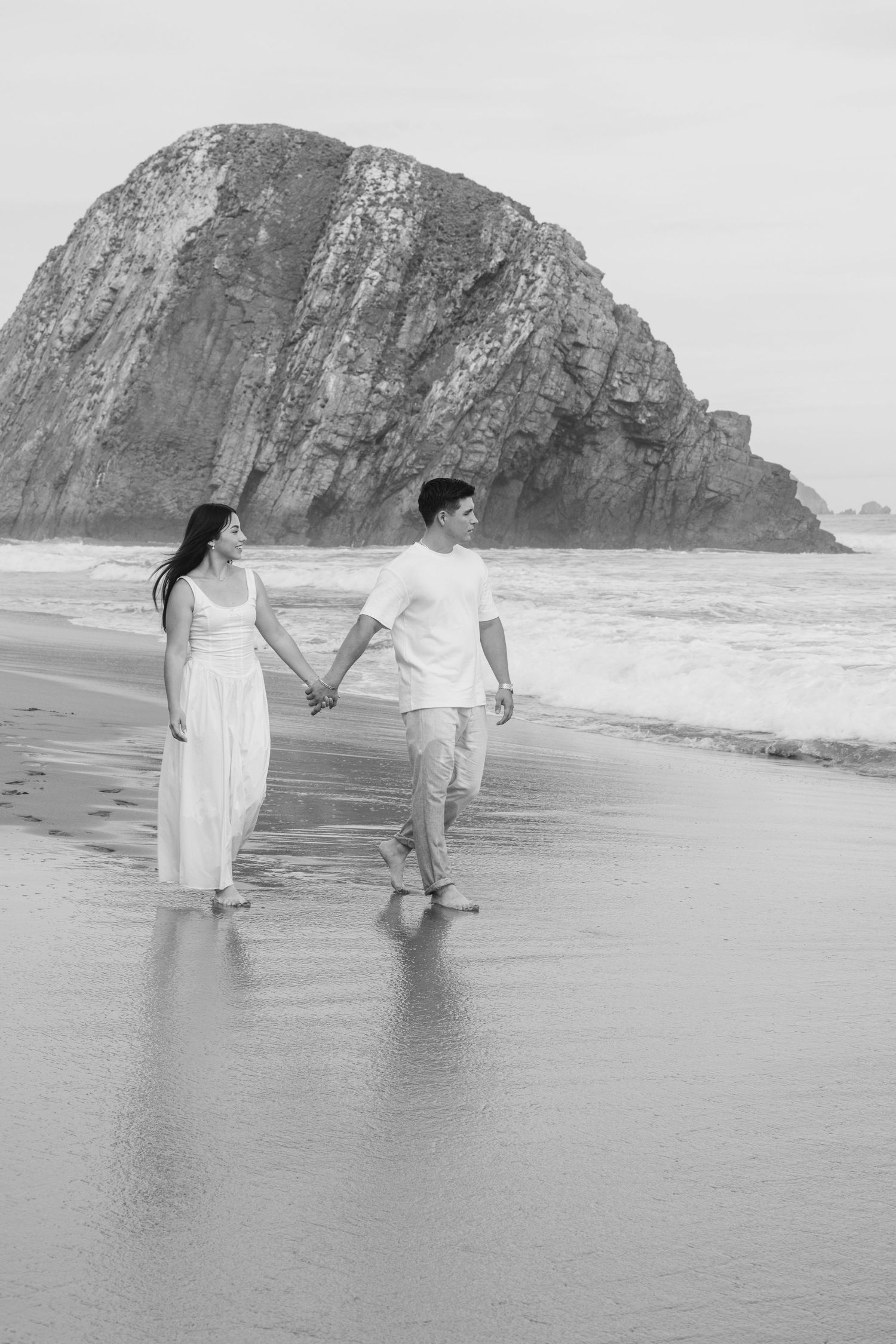 Couple holding hands walking barefoot on beach in black and white, large rock formation and ocean waves in background.