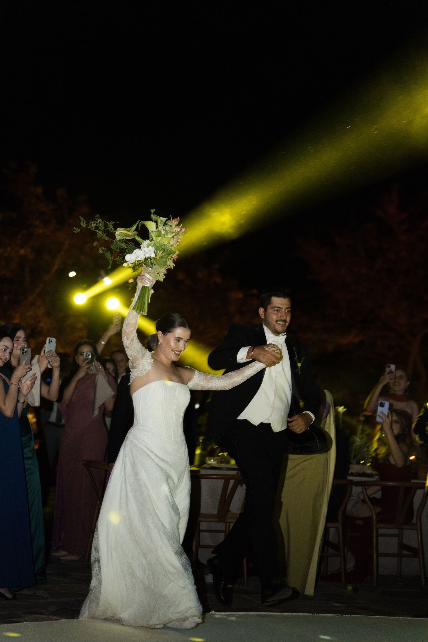 Bride and groom making grand entrance at night wedding reception with spotlight and bouquet raised.