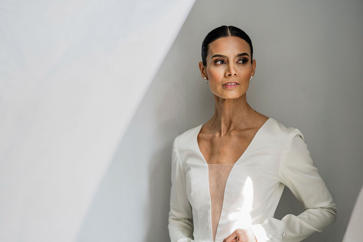 Bride in minimalist deep V-neck white long-sleeve gown poses confidently against a light gray wall.