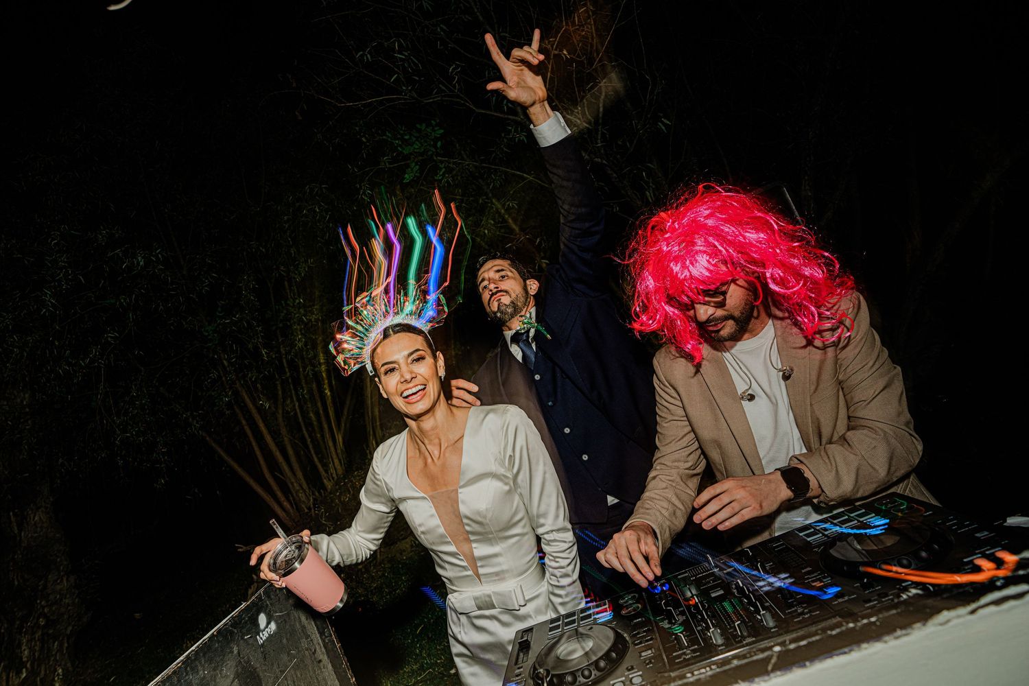 Bride wearing colorful crown headpiece laughs with DJ in pink wig at lively wedding reception party.