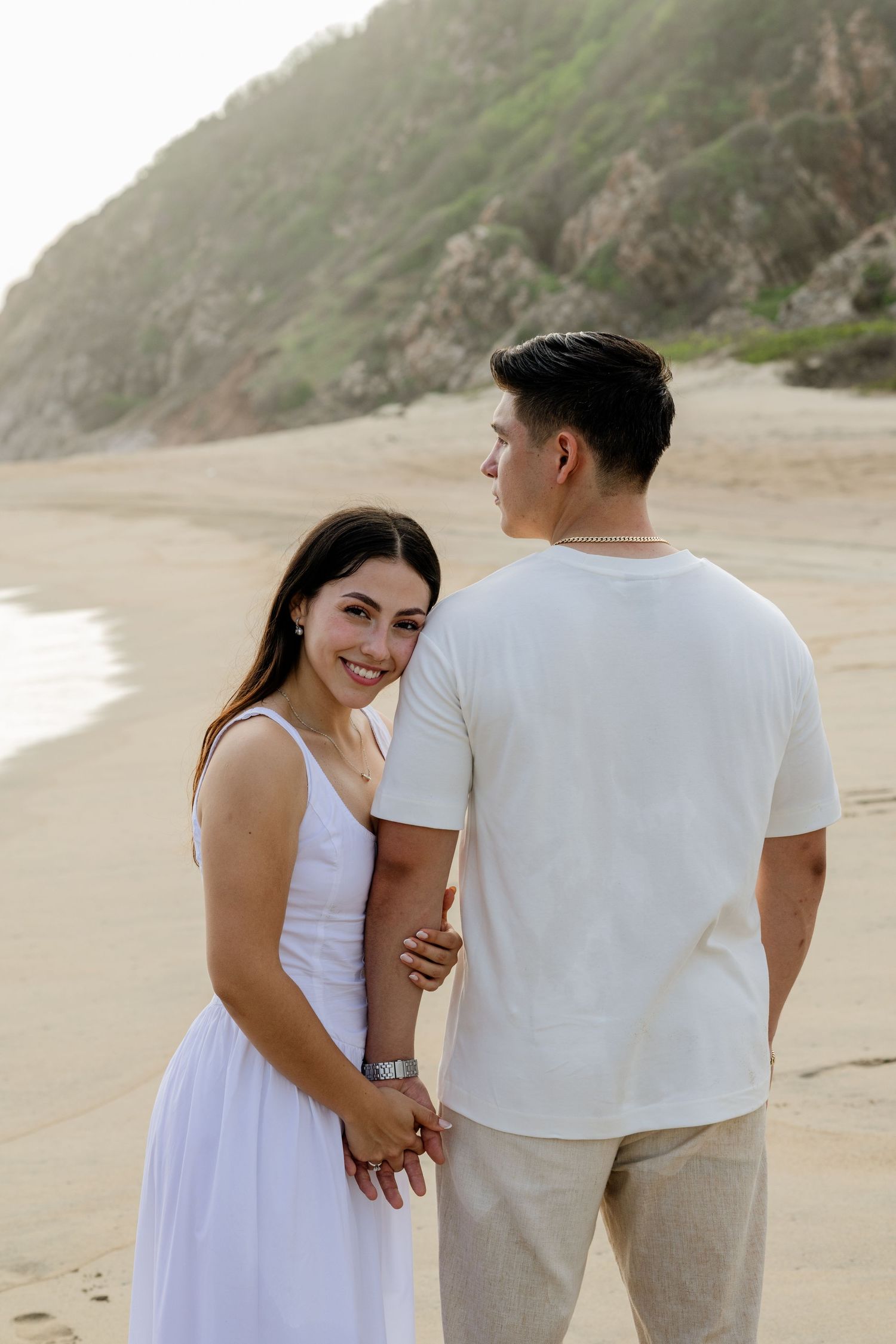 Couple holding hands on a sandy beach, woman in white dress smiling at camera, man in white shirt facing away.