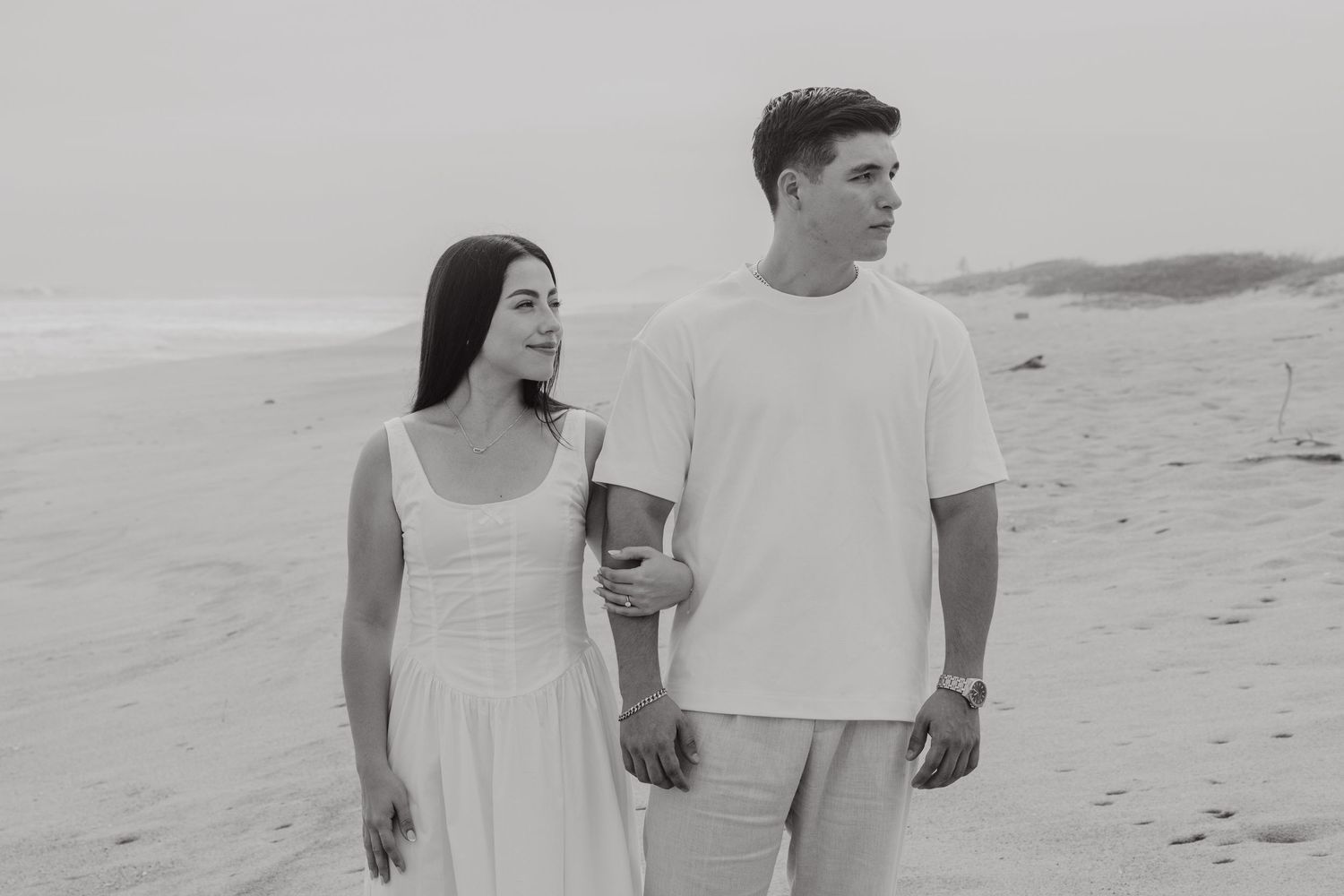 Black and white photo of a couple walking on a beach, woman in white dress holding man's arm, ocean in background.