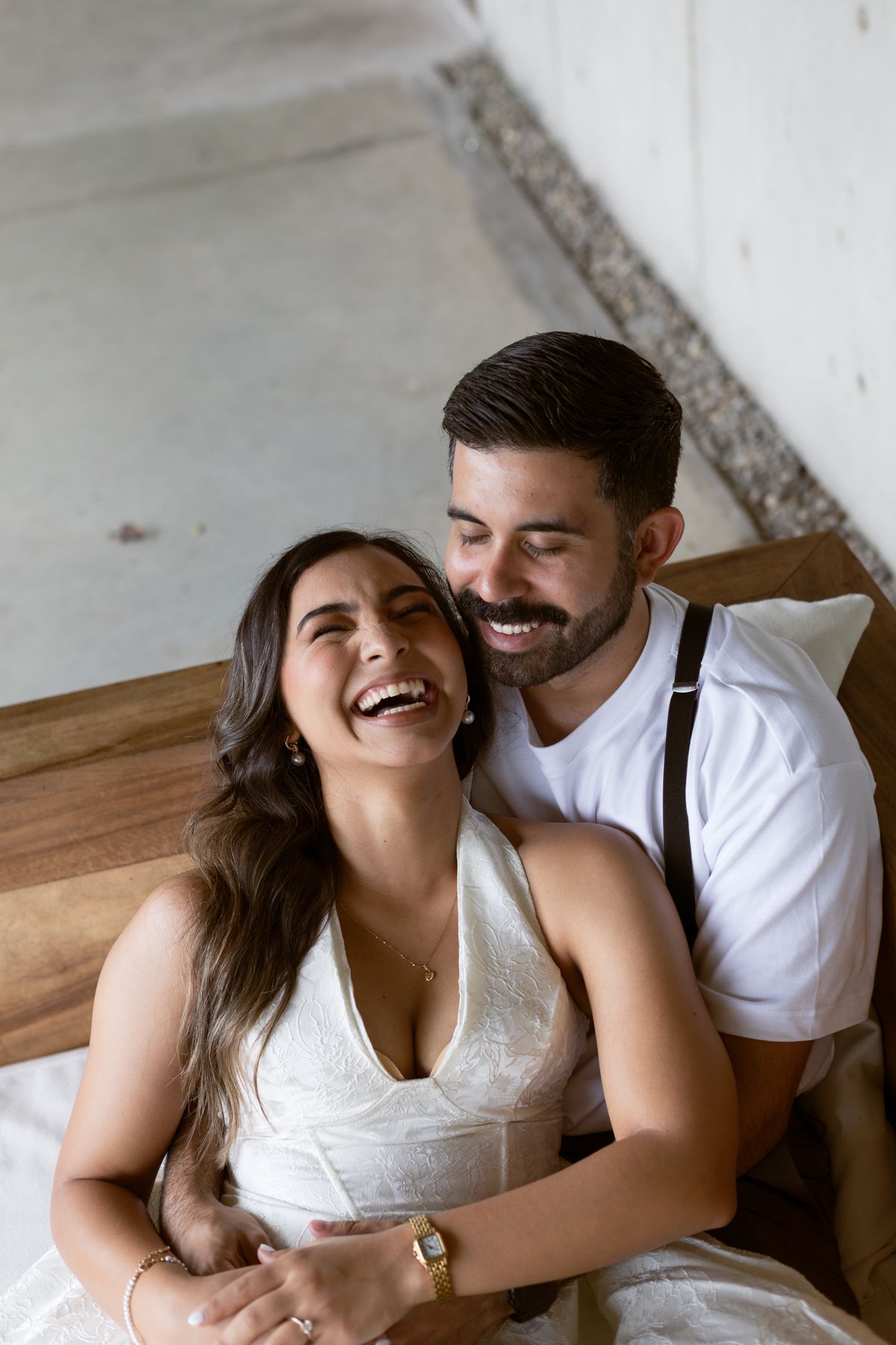 Joyful couple laughing together on a bed, bride in white dress embraced by bearded groom in white shirt.