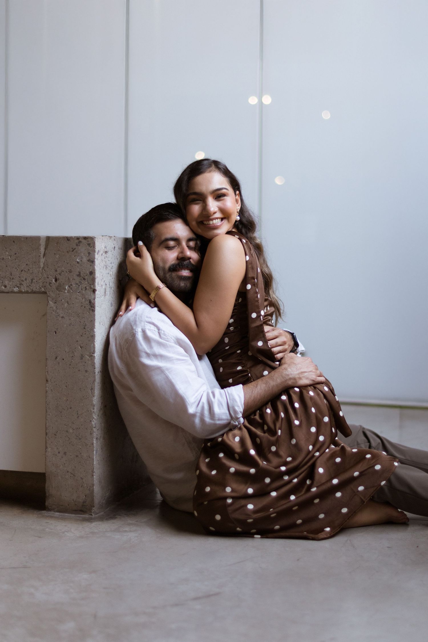 Happy couple sitting on floor, woman in brown polka dot dress embracing man in white shirt against wall.