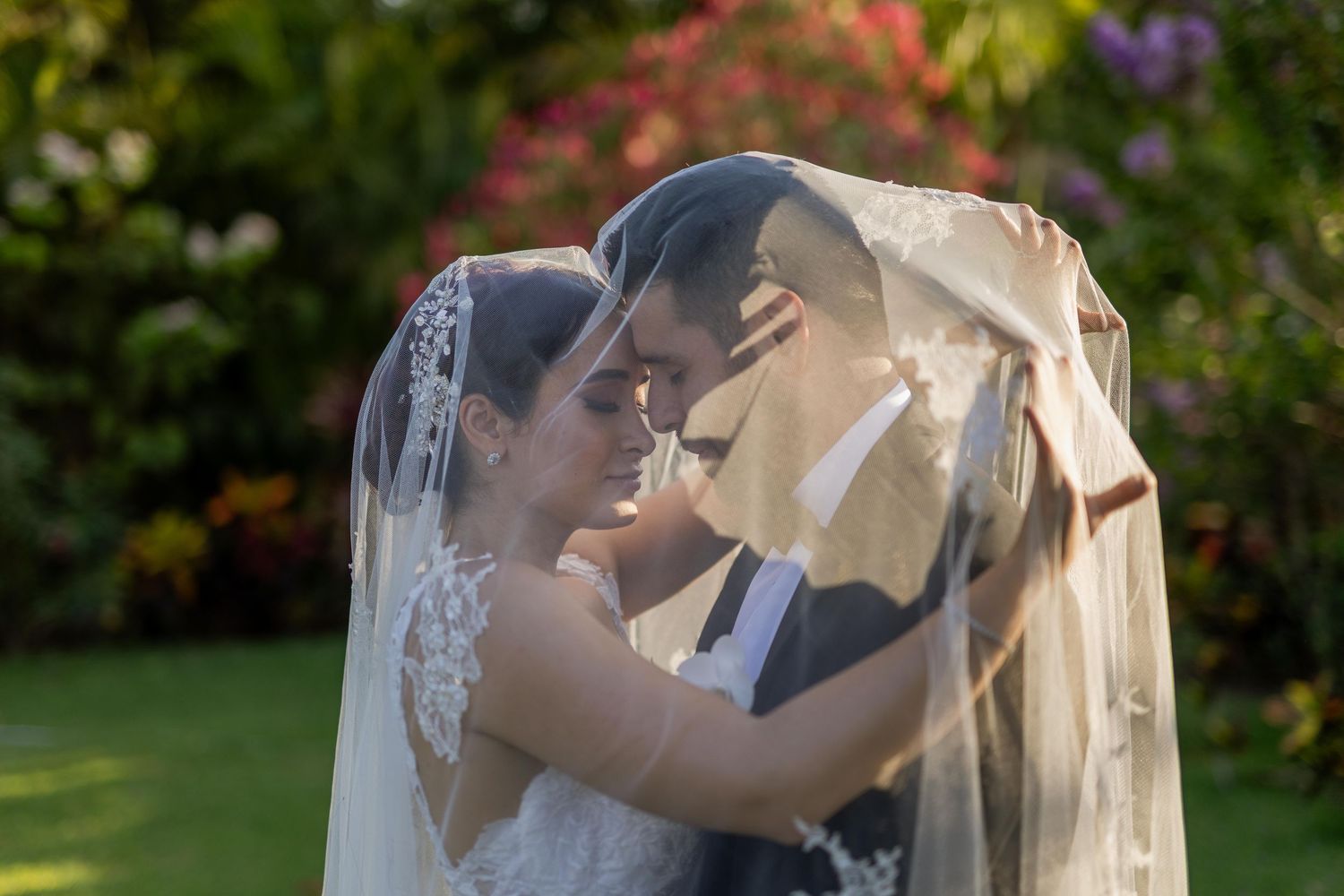 Romantic couple shares an intimate moment beneath a flowing bridal veil surrounded by lush garden greenery.